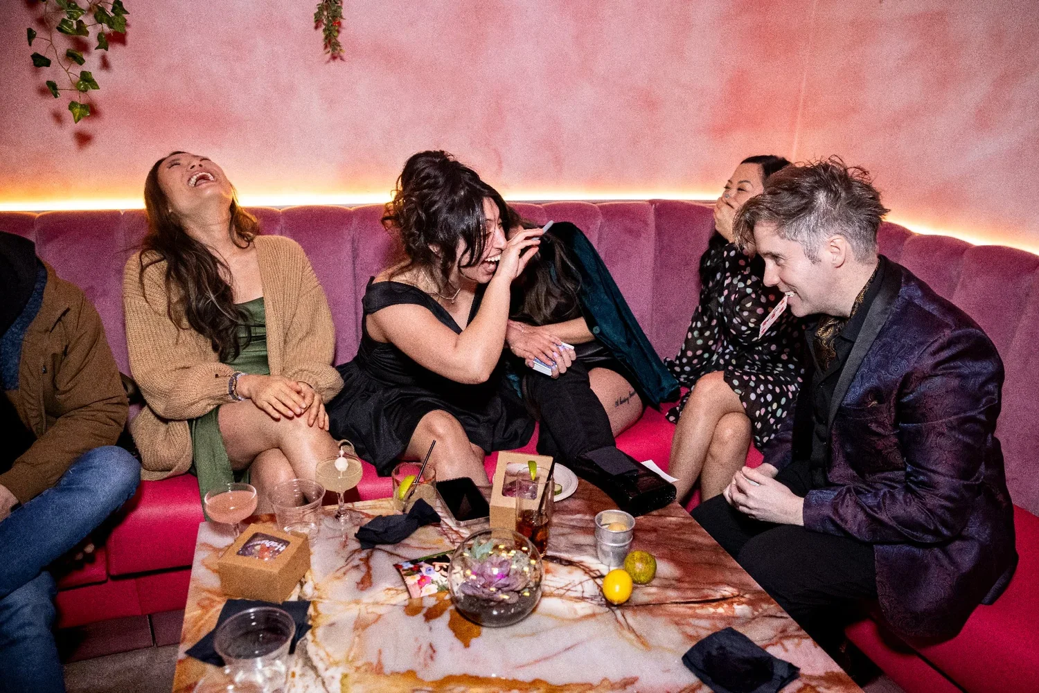Group of friends laughing and enjoying drinks at a party in a pink-themed lounge.