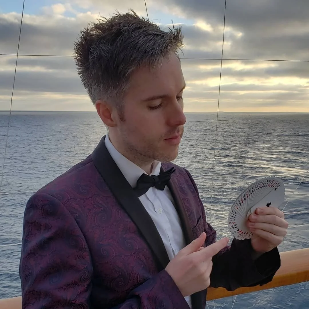 Man in a purple patterned tuxedo with a black bow tie, holding a fanned deck of playing cards, standing on a boat with ocean and cloudy sky in the background.