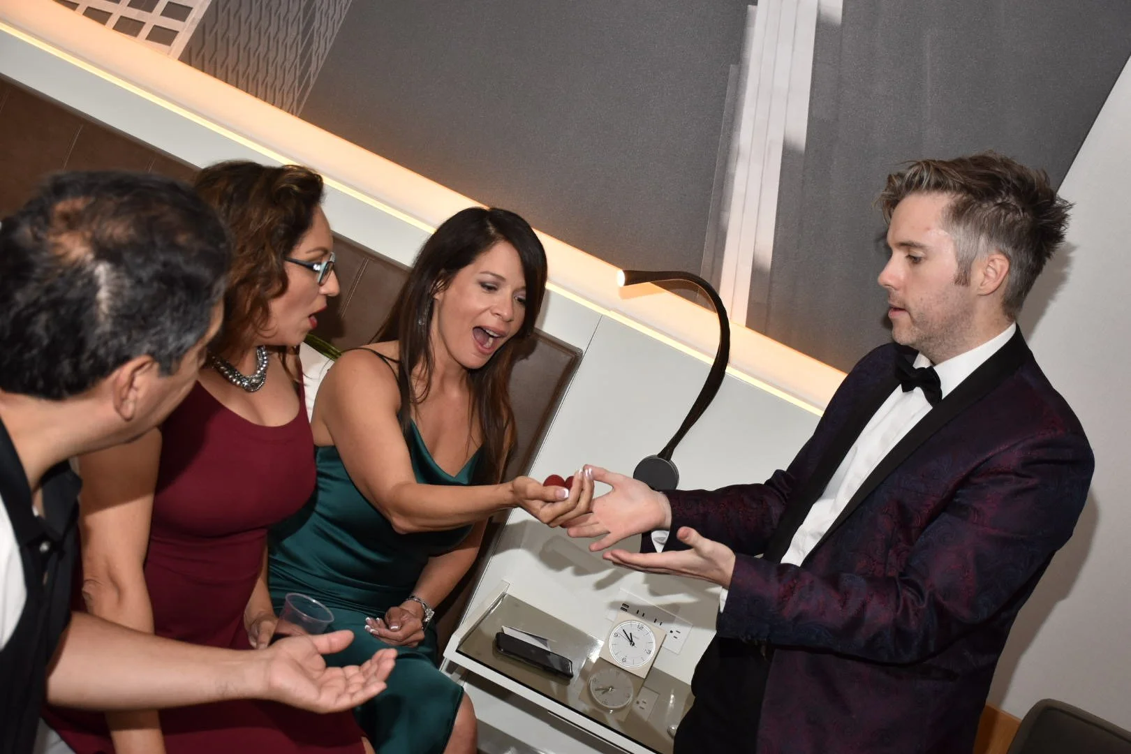 A woman in a green dress is receiving a ring from a man in a tuxedo at a formal event. Three other people are watching, one holding a glass, in a modern, well-lit room.