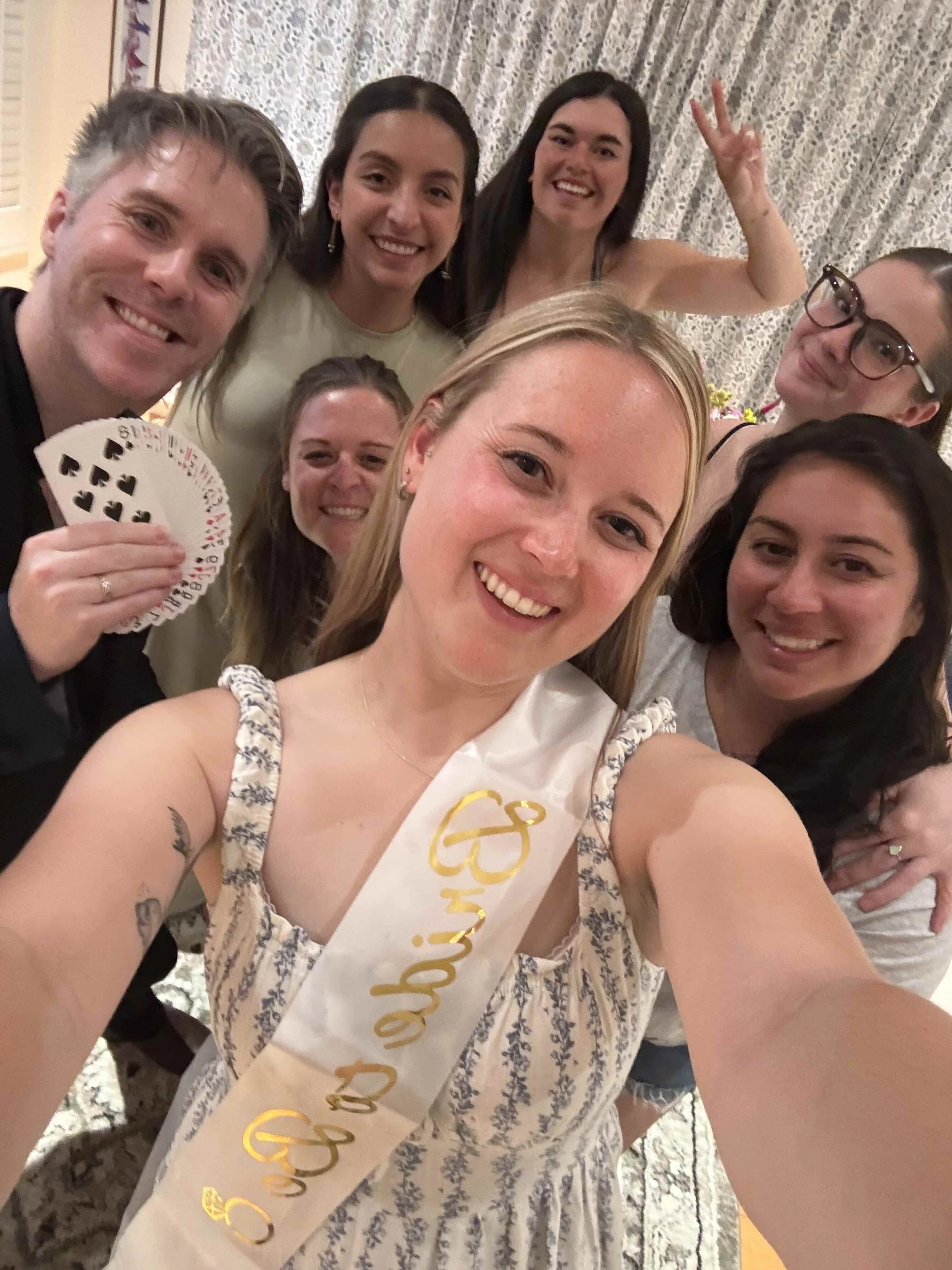 Group of women celebrating at a party, with one woman wearing a sash that says 'Glam' and holding playing cards, smiling at the camera.