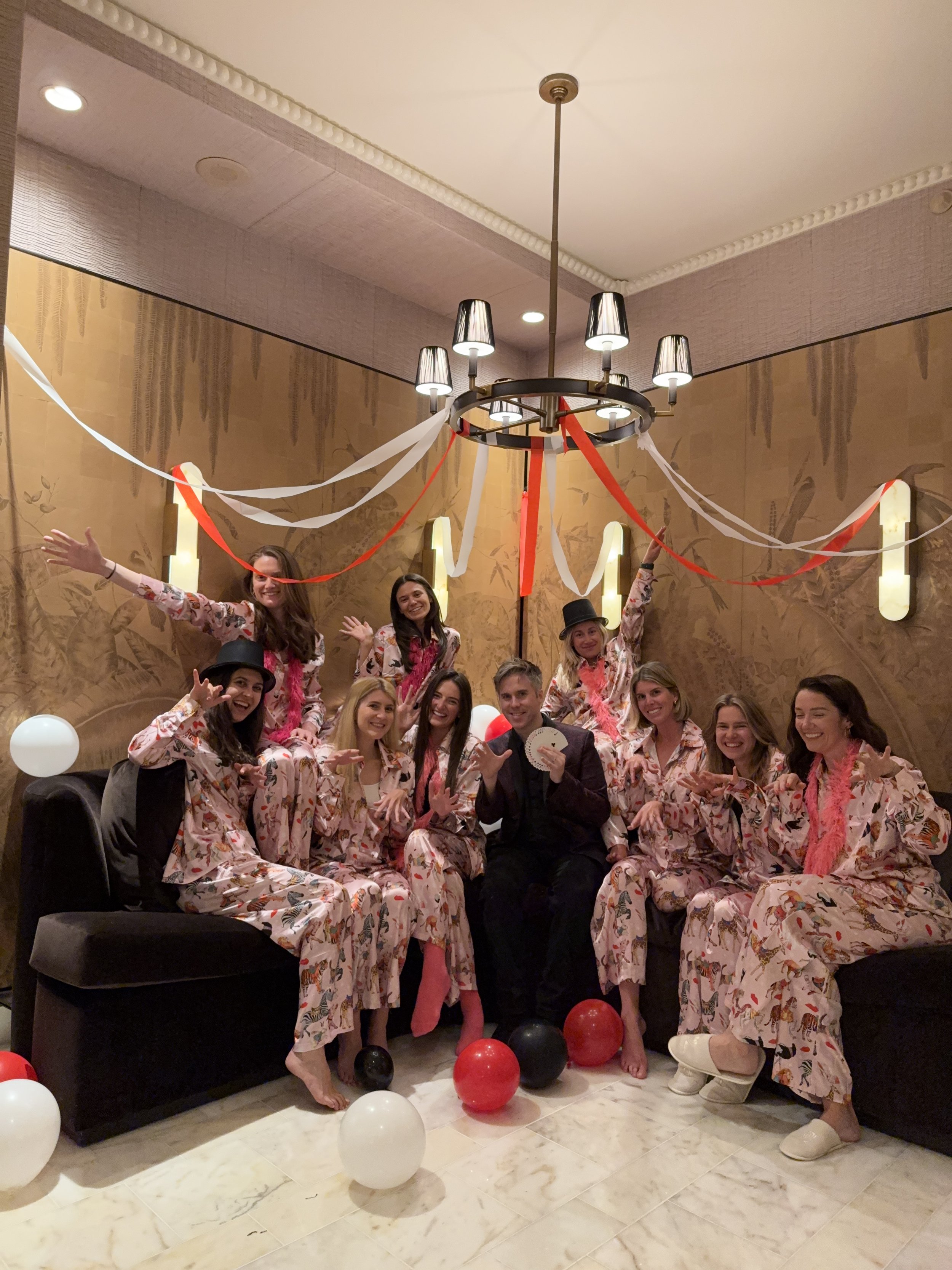 Group of people in matching pajamas celebrating in a living room with balloons and streamers, with a chandelier overhead.