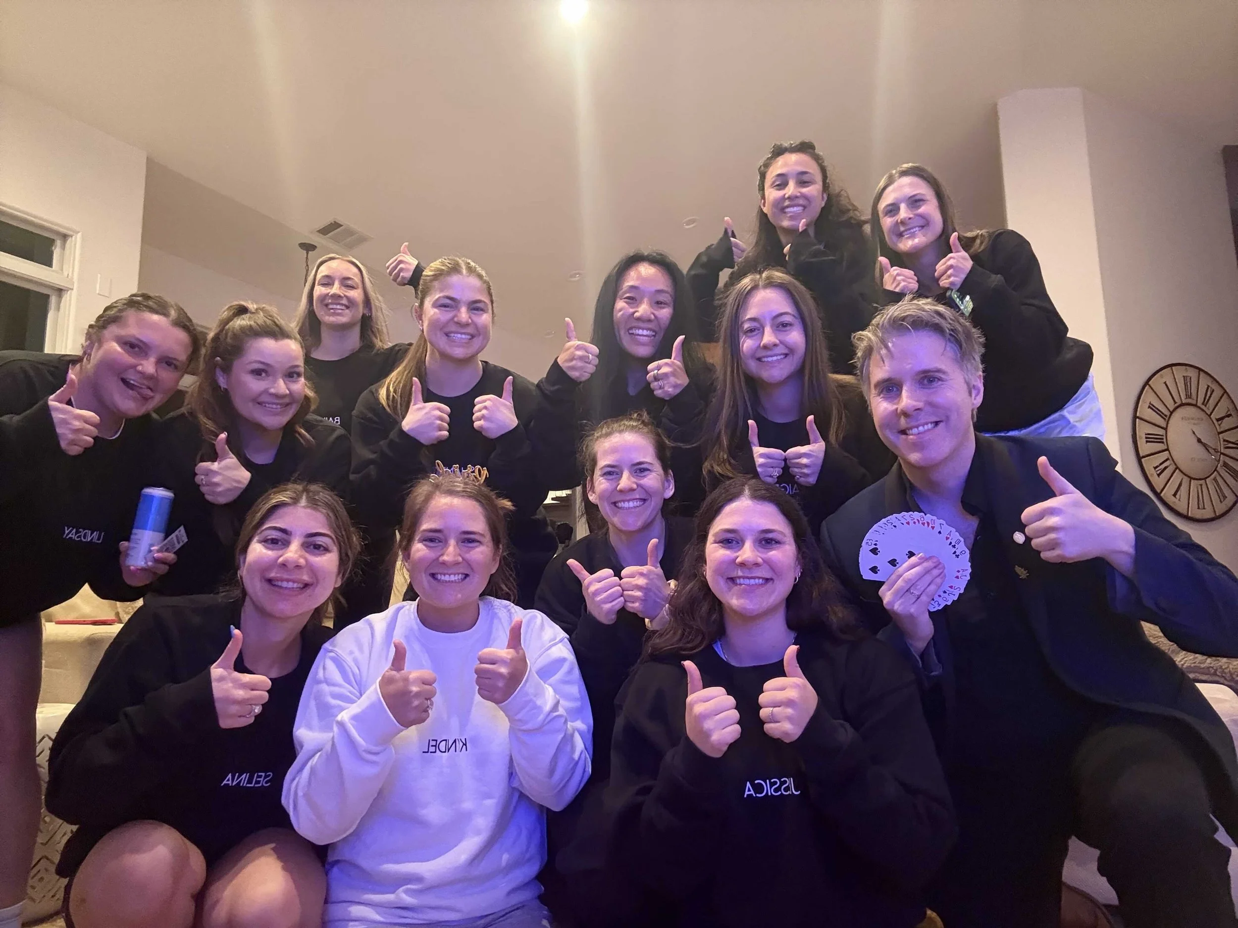 Group of young women and a man smiling and giving thumbs up in a living room, celebrating a birthday with a deck of cards and a stuffed animal on one person's lap.