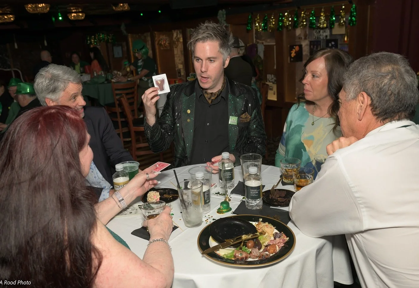 A group of six adults sitting around a table at a party or celebration, with one man in the center holding a card and engaging others in a game, decorated with green and gold festive ornaments.