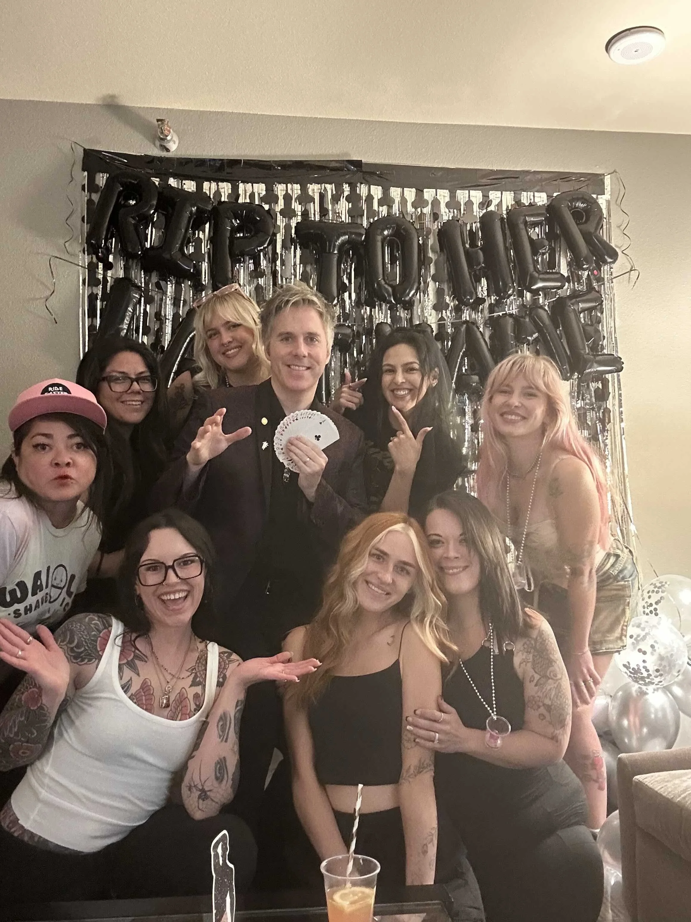 Group of people celebrating at a party with black "HAPPY BIRTHDAY" balloons and balloons on the right side of the image.