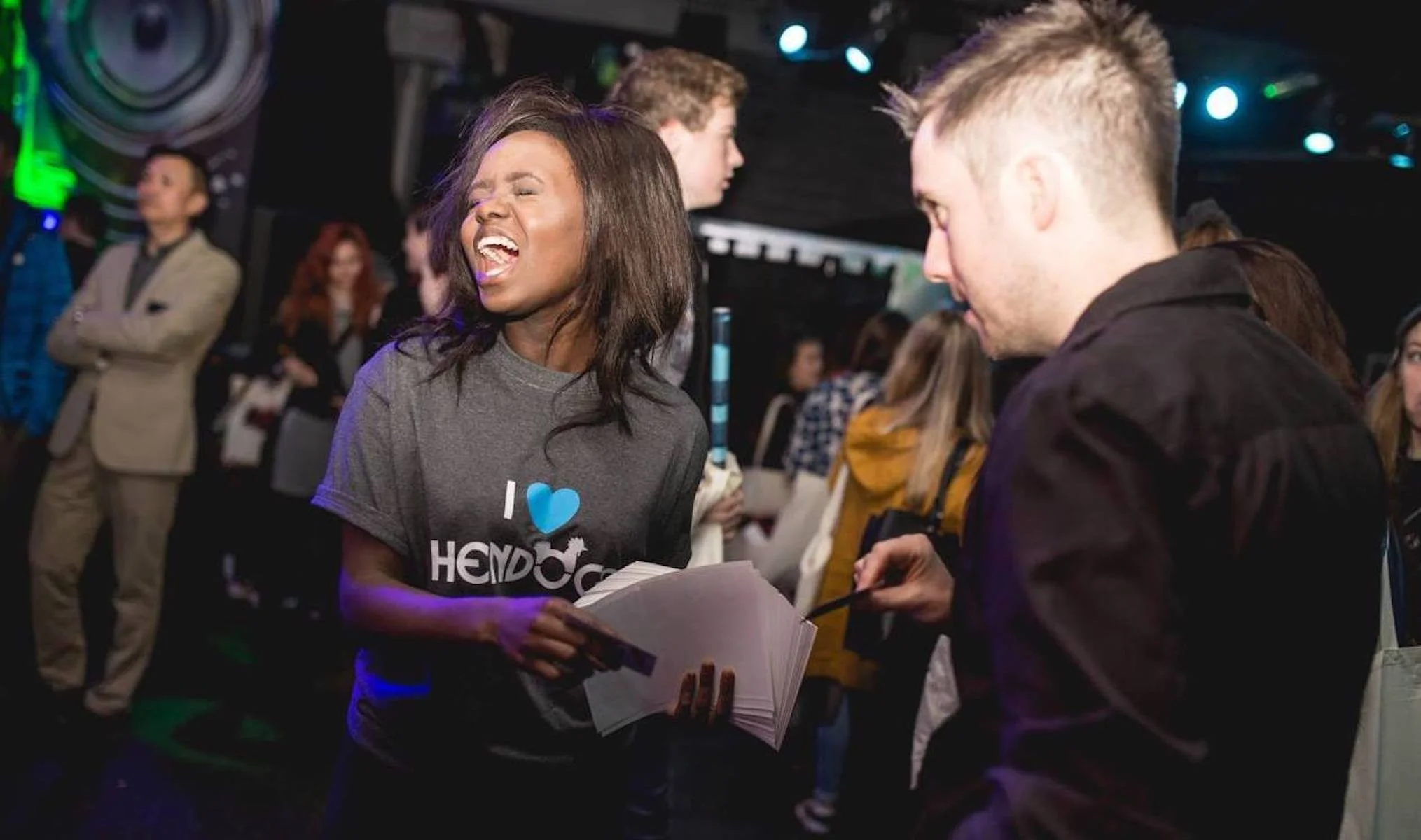 A woman with long dark hair smiling and singing at a nightclub or party, holding a booklet, surrounded by other people in a dimly lit environment with colorful lights.