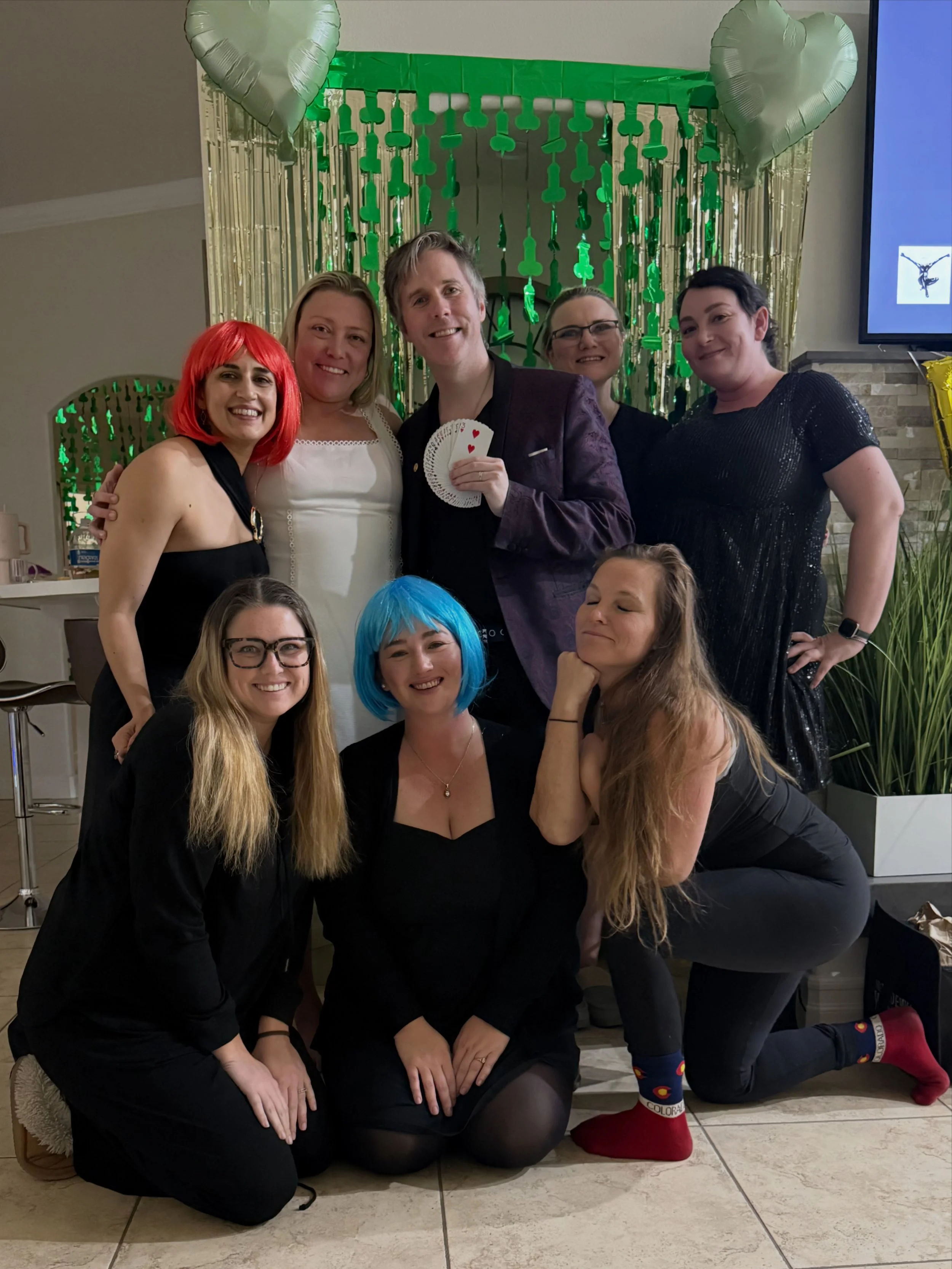 Group of eight friends celebrating at a party, with colorful balloons, gold fringe curtains, and a large blue screen in the background. Some are wearing wigs and festive clothing, smiling and posing together.