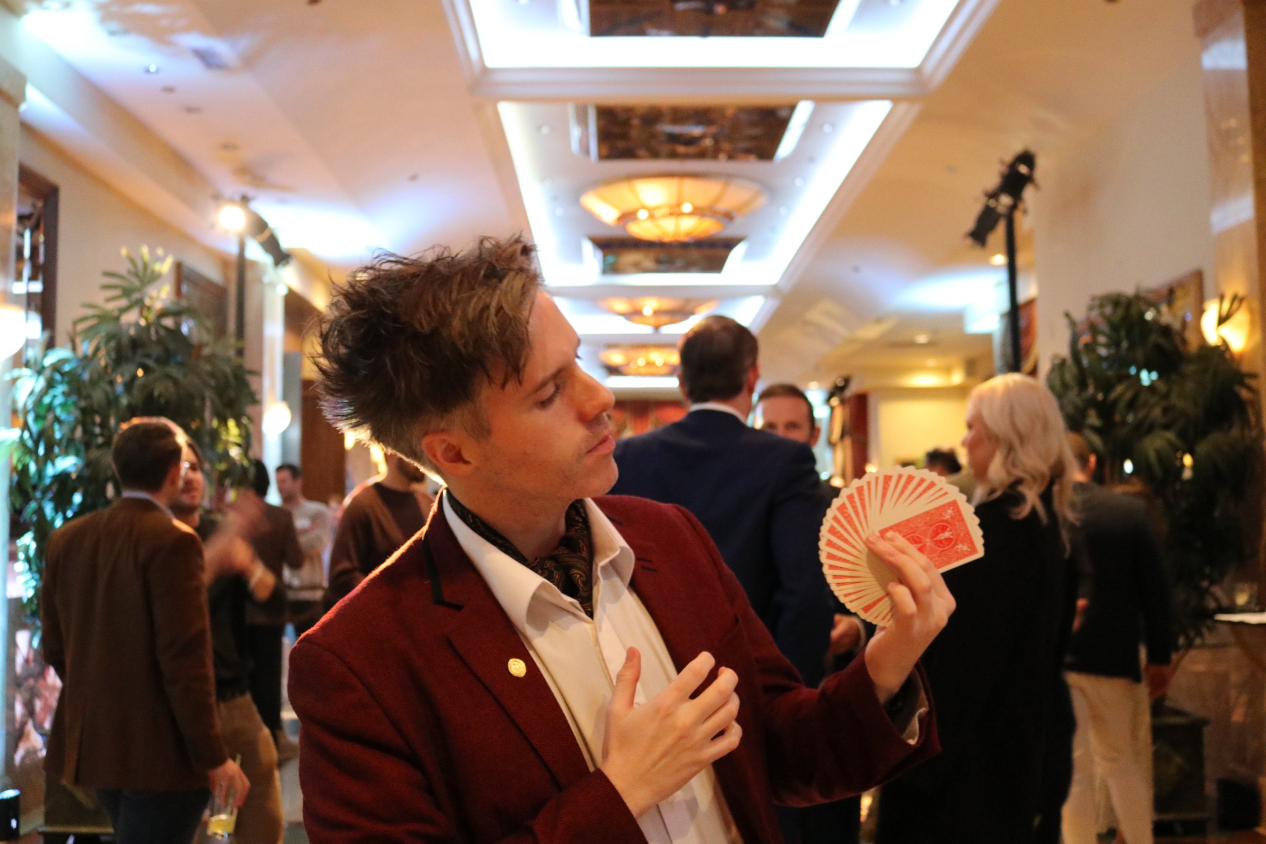 A young man dressed in a tuxedo with a red jacket and a patterned tie is playing cards in a lively, well-lit party or casino setting. He appears focused and is holding a fan of red playing cards in his right hand. The background shows other people mingling and bright headers with lights, greenery, and elegant decor.