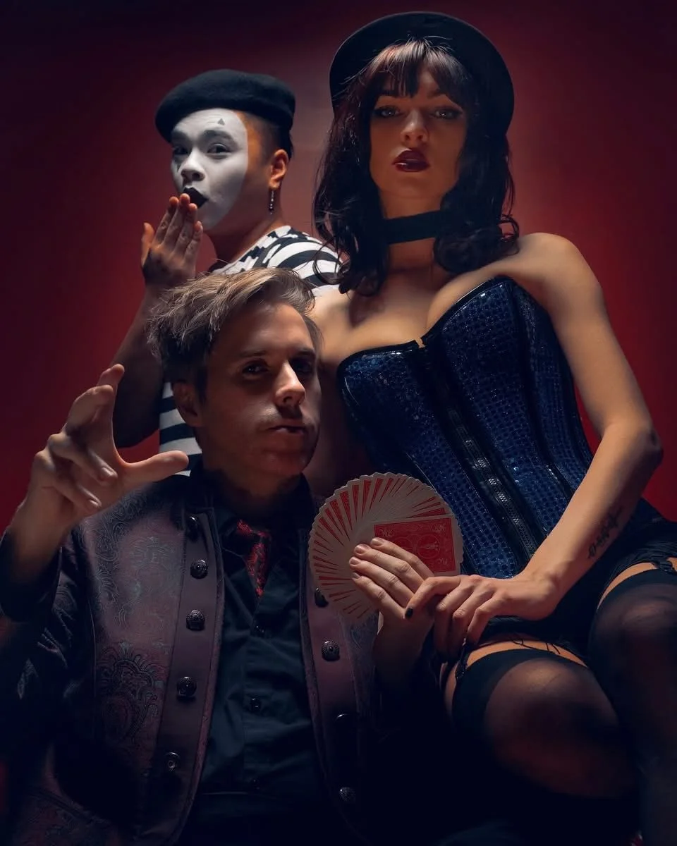 Three performers dressed in vintage and theatrical costumes pose against a red background, including a woman in a corset and hat, a man with a fan, and a mime in makeup and striped shirt.
