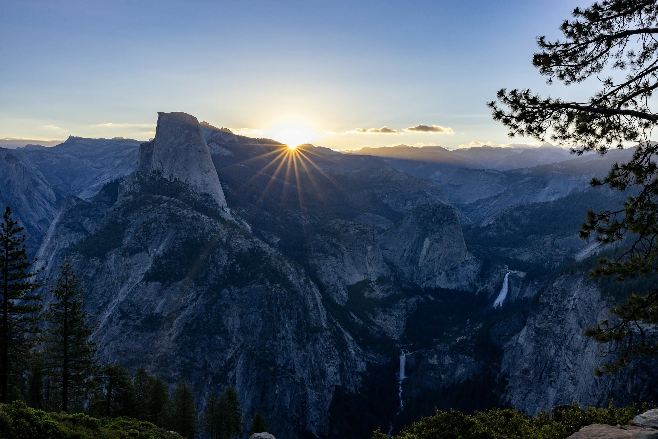 Sunset over rugged mountain landscape with waterfalls and pine trees in foreground.
