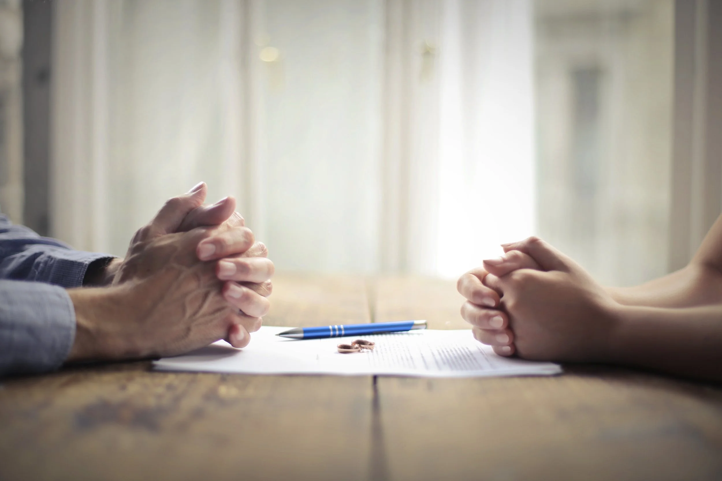 Close-up of two pairs of hands lying on a wooden table, clasped together. On the table, there is a blue pen and some papers. The background shows a bright, softly lit room with curtains.