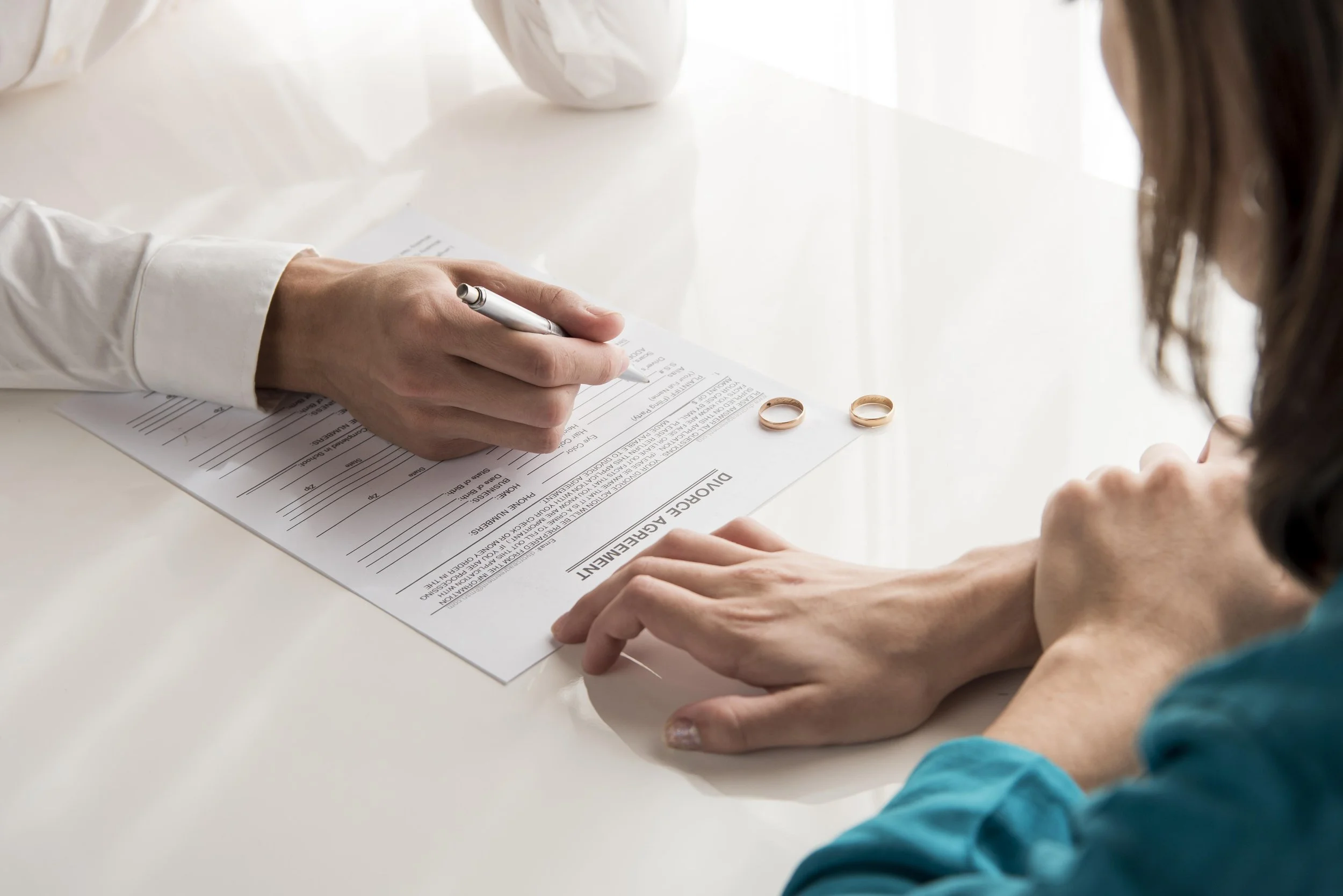 Close-up of a person signing a document titled 'DURALIST AGREEMENT' with wedding rings placed on the paper, indicating a marriage or legal agreement, beside another person with long hair.