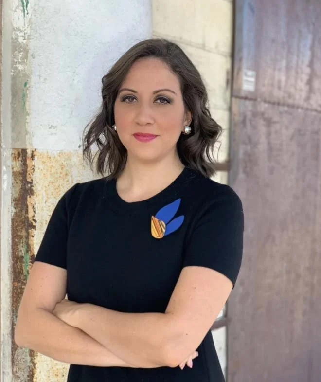 Woman with shoulder-length wavy brown hair, wearing pearl earrings and a black shirt with a blue and orange pin, standing outdoors near a weathered white-painted pillar.