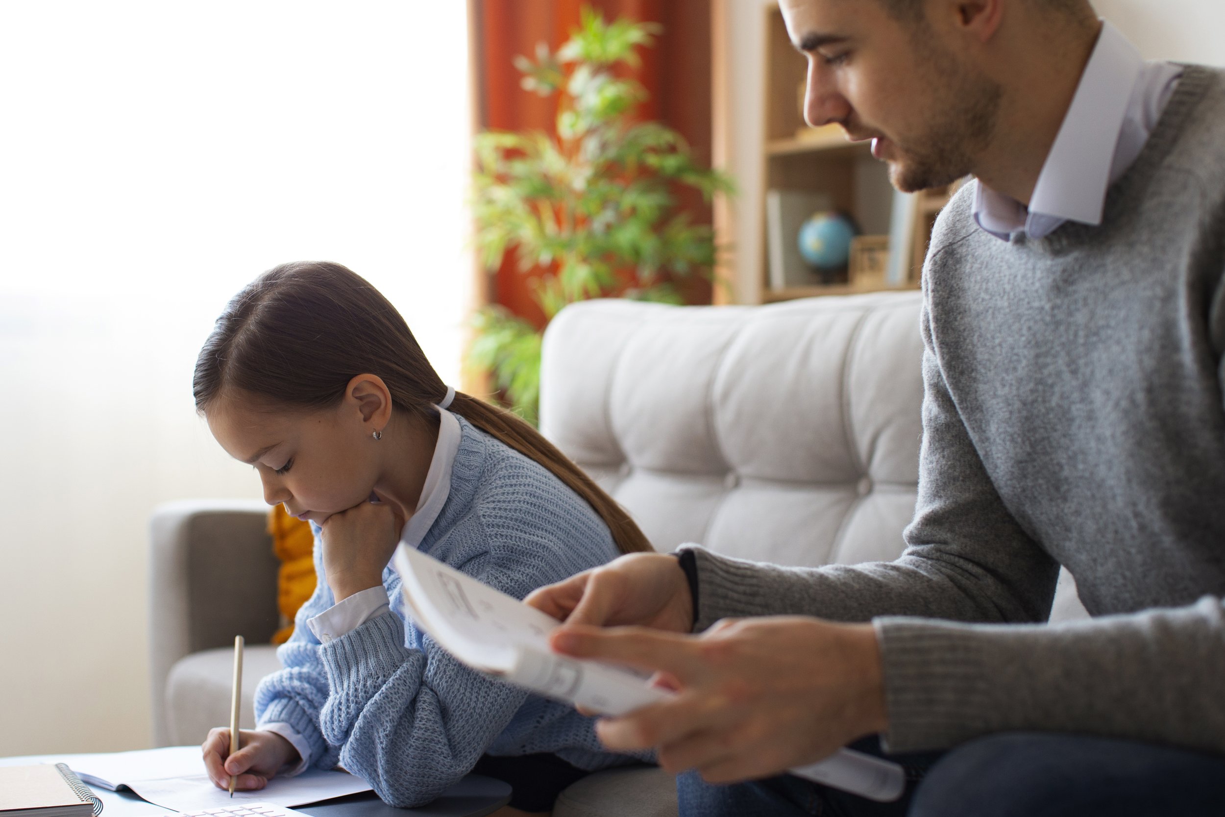 A young girl with brown hair sitting on a beige couch, writing in a notebook with a pencil. An adult man, possibly her father, sitting next to her, holding a book or magazine, and looking down at her. Both are in a well-lit room with a bookshelf, a globe, and a potted plant in the background.