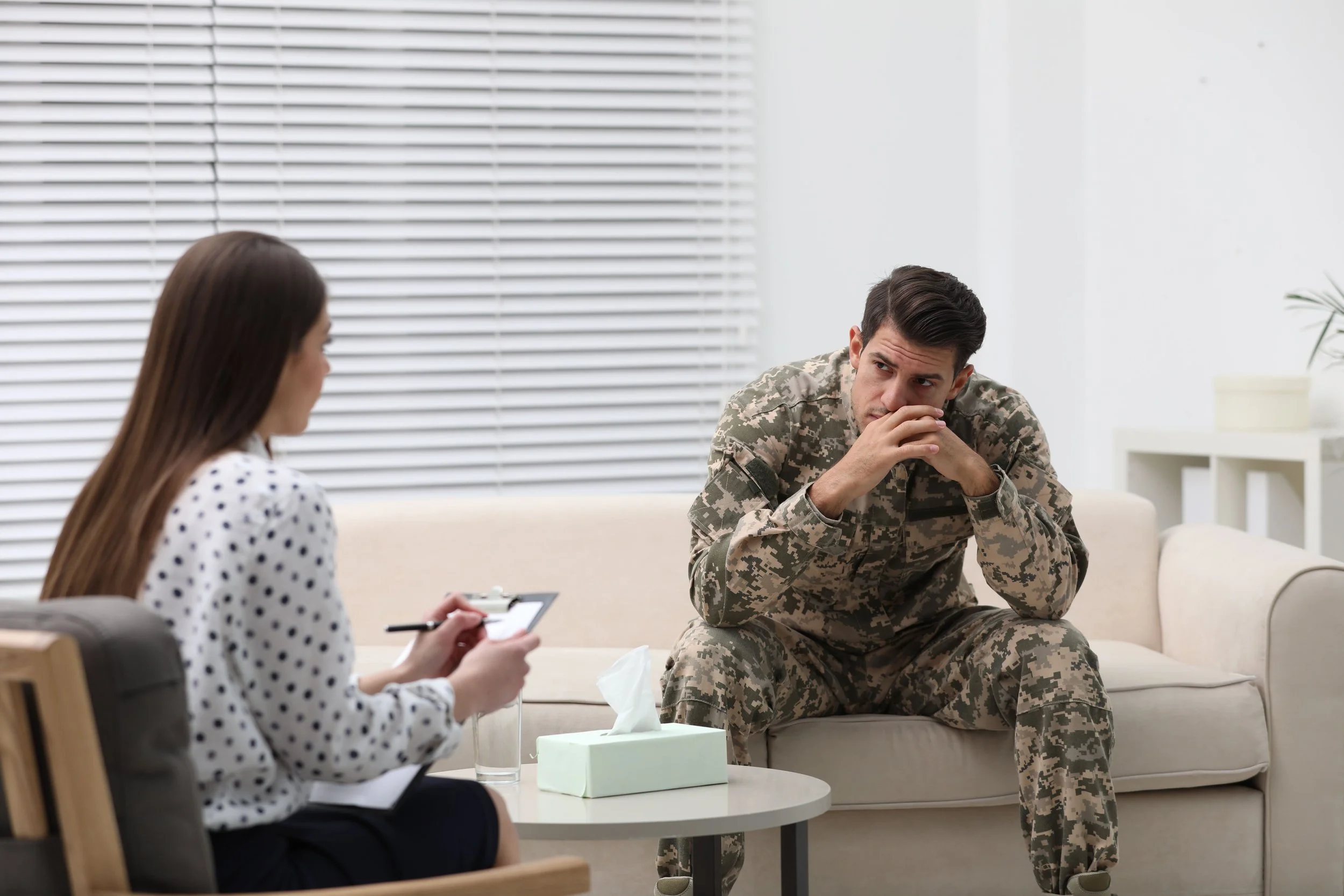 A man in military uniform sitting on a beige couch, appearing deep in thought. A woman in a polka dot blouse, taking notes, is seated across from him in a consultation or therapy session. The room has white walls, a window with horizontal blinds, and a small white table with tissues and a glass of water.