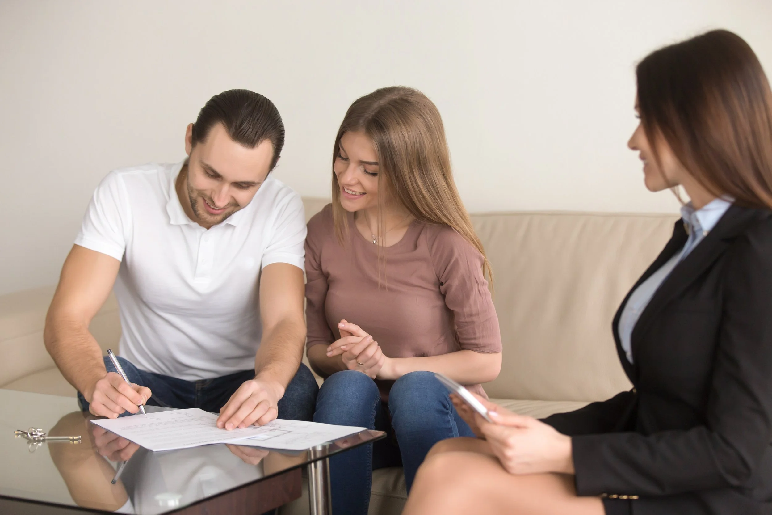 Three people, two women and one man, are sitting together in a professional setting. They are reviewing a document on a glass table, with the man holding a pen. It appears to be a meeting or consultation, with a neutral background and a relaxed, engaged atmosphere.