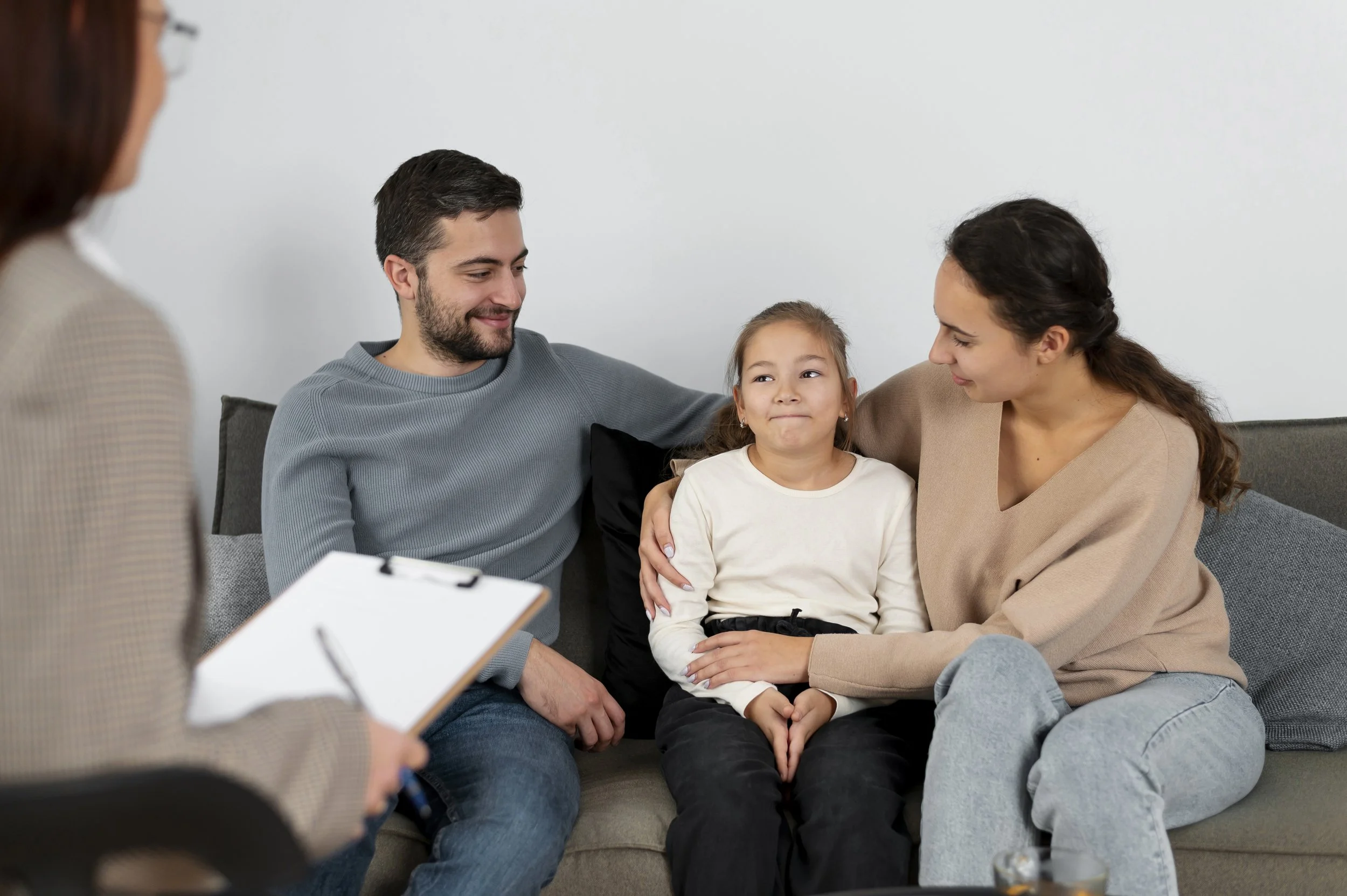 A family consisting of a young girl, a man, and a woman sitting on a grey couch during a therapy or counseling session. They are engaging with a person holding a clipboard, with a mostly blank white wall in the background.