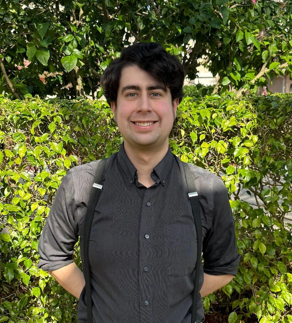A young man with dark, curly hair smiling, standing outdoors in front of green bushes and trees, wearing a dark button-up shirt and carrying a backpack.