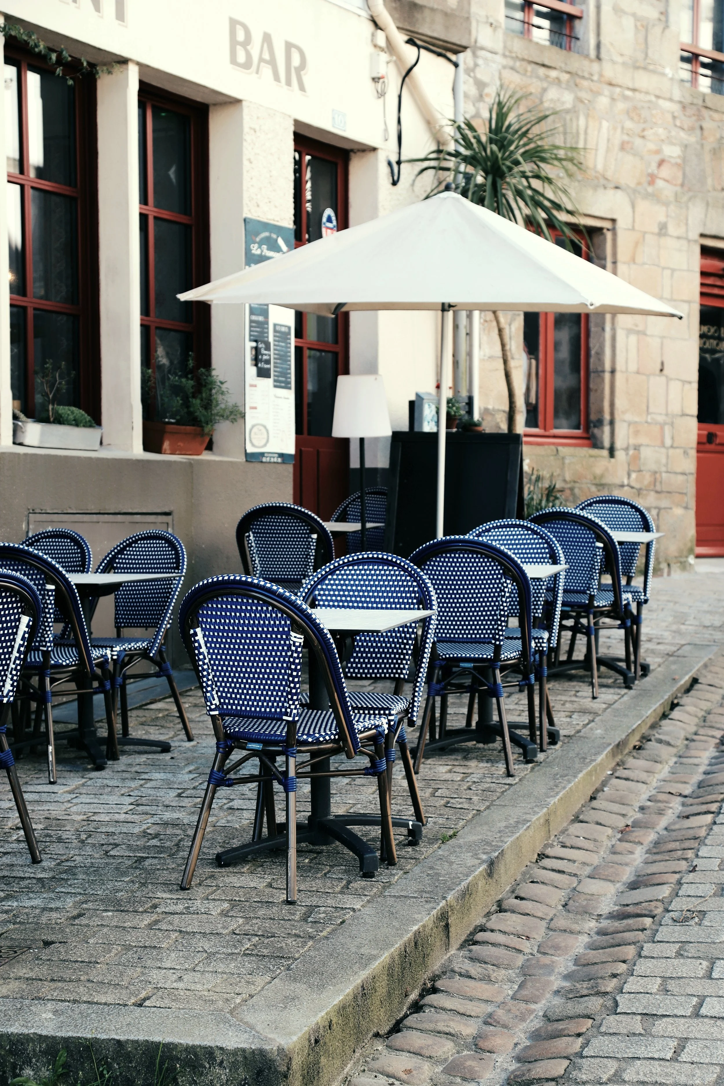 Terrasse de café avec plusieurs tables et chaises en osier en face d'un bar, sous un grand parasol blanc, située sur une rue pavée