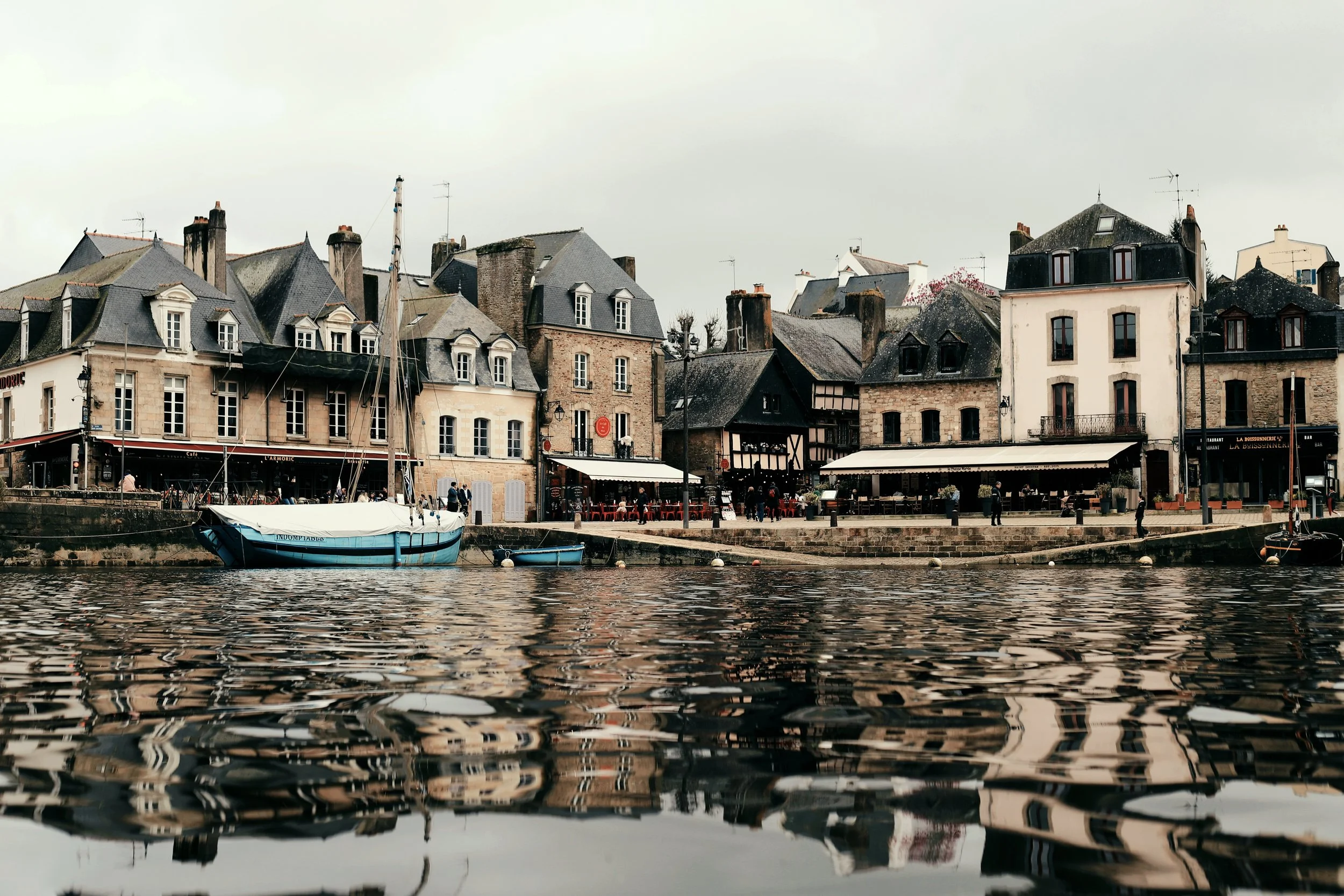 Vue d'un port avec des maisons anciennes en pierre, un bateau à voile et des petits bateaux en eaux calmes, reflet des bâtiments dans l'eau, ciel nuageux.