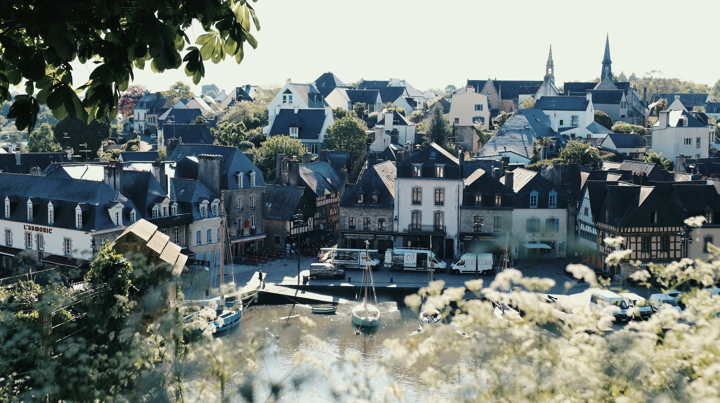 Vue d'un village côtier avec des maisons anciennes, un port avec des bateaux, et des églises en arrière-plan, encadré par des feuilles d'arbres.