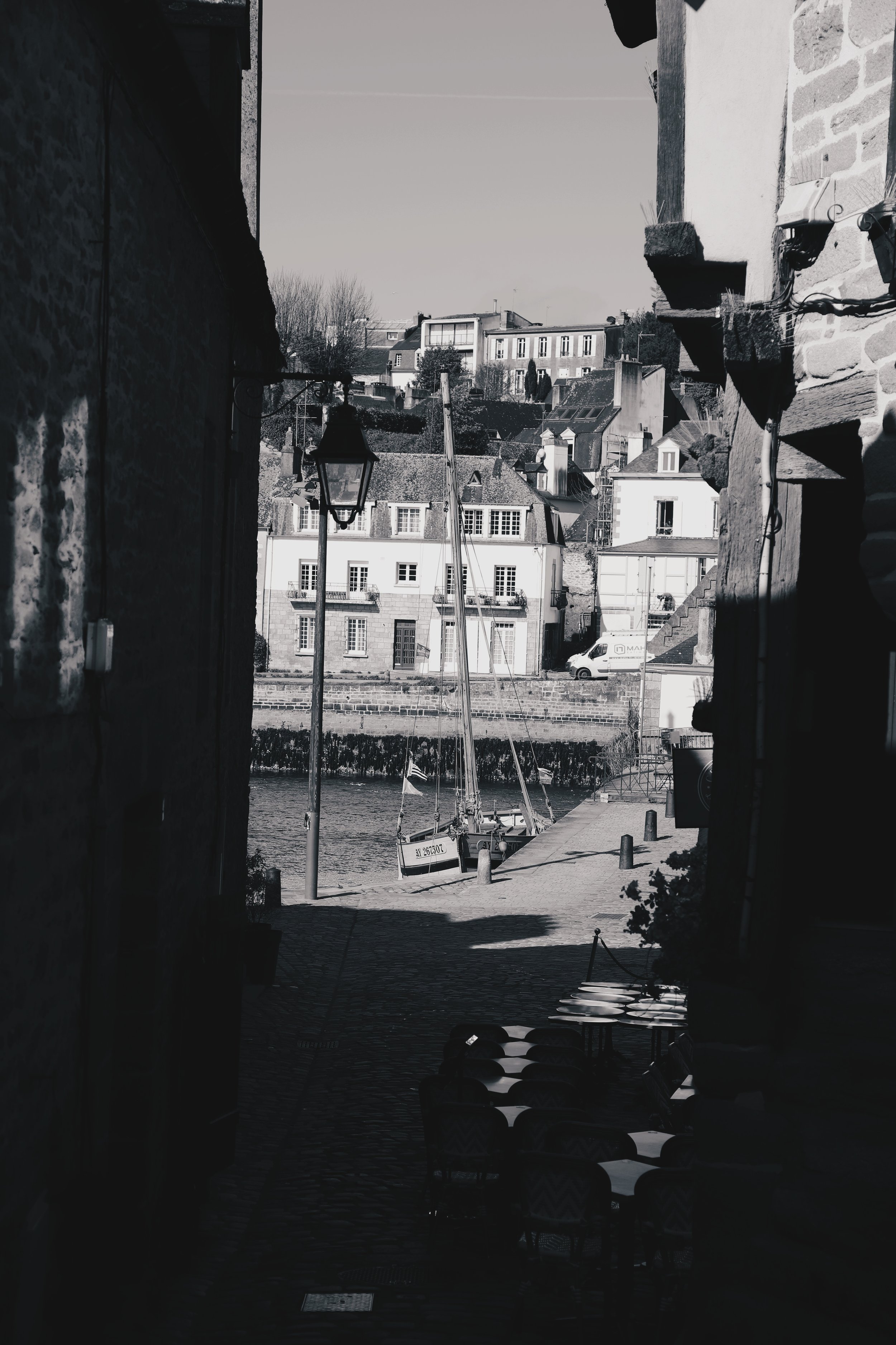 Vue d'un port avec un bateau amarré, des maisons en arrière-plan, un lampadaire et des tables dans une rue étroite en style noir et blanc.