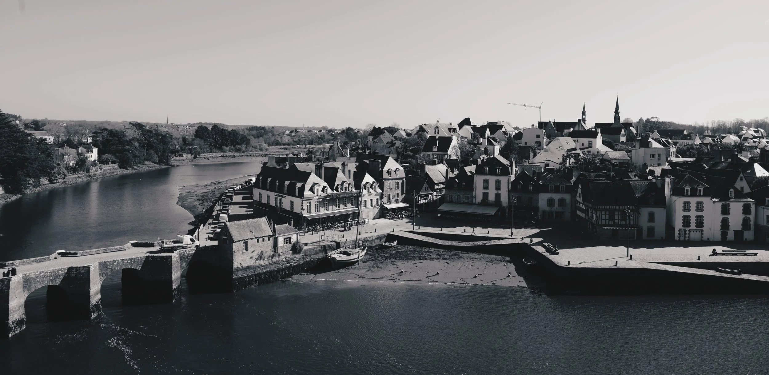 Vue d'une ville au bord d'une rivière avec des bâtiments historiques et une église avec des flèches pointues, en noir et blanc.