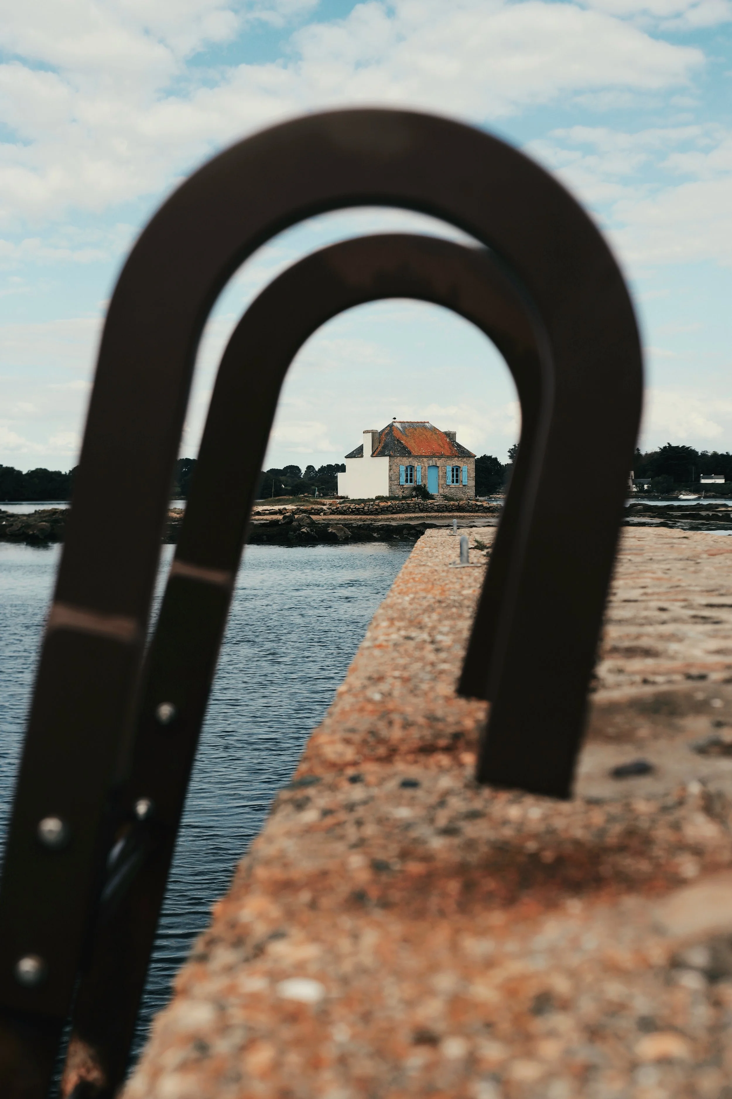 Une cabane blanche avec volets bleus, vue à travers une structure en métal, située sur une petite île ou un rivage, avec un ciel partiellement nuageux.