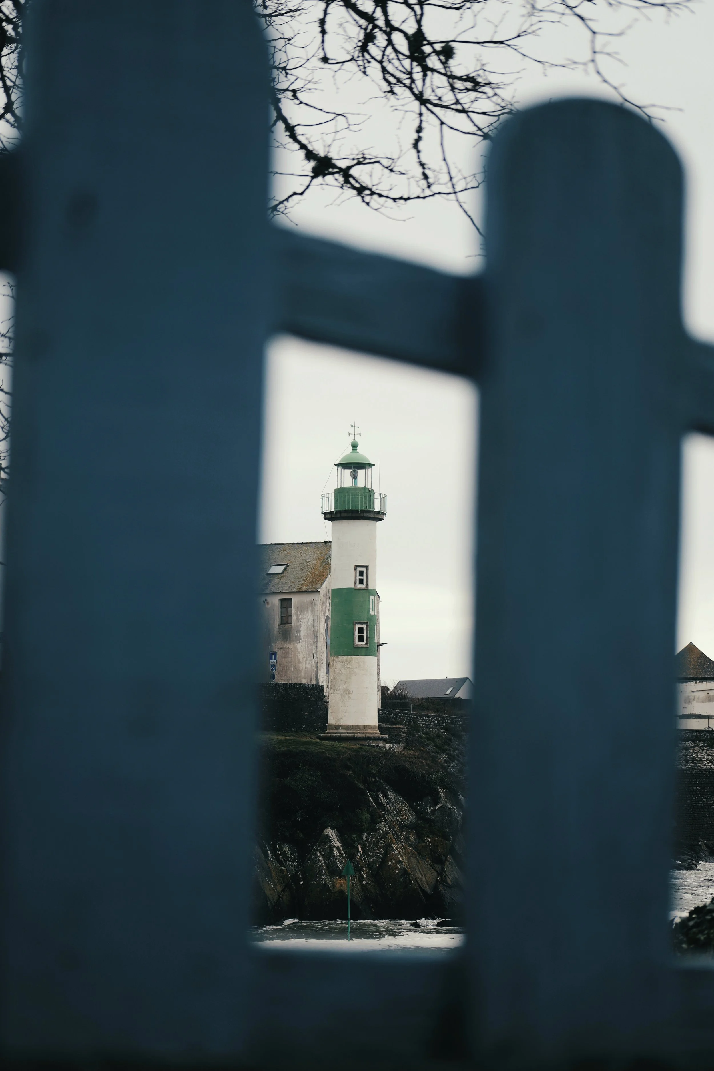 Phare ancien avec une tour en béton blanche et une coiffe verte, vu à travers une clôture en bois, par temps gris, avec des arbres dénudés en arrière-plan.