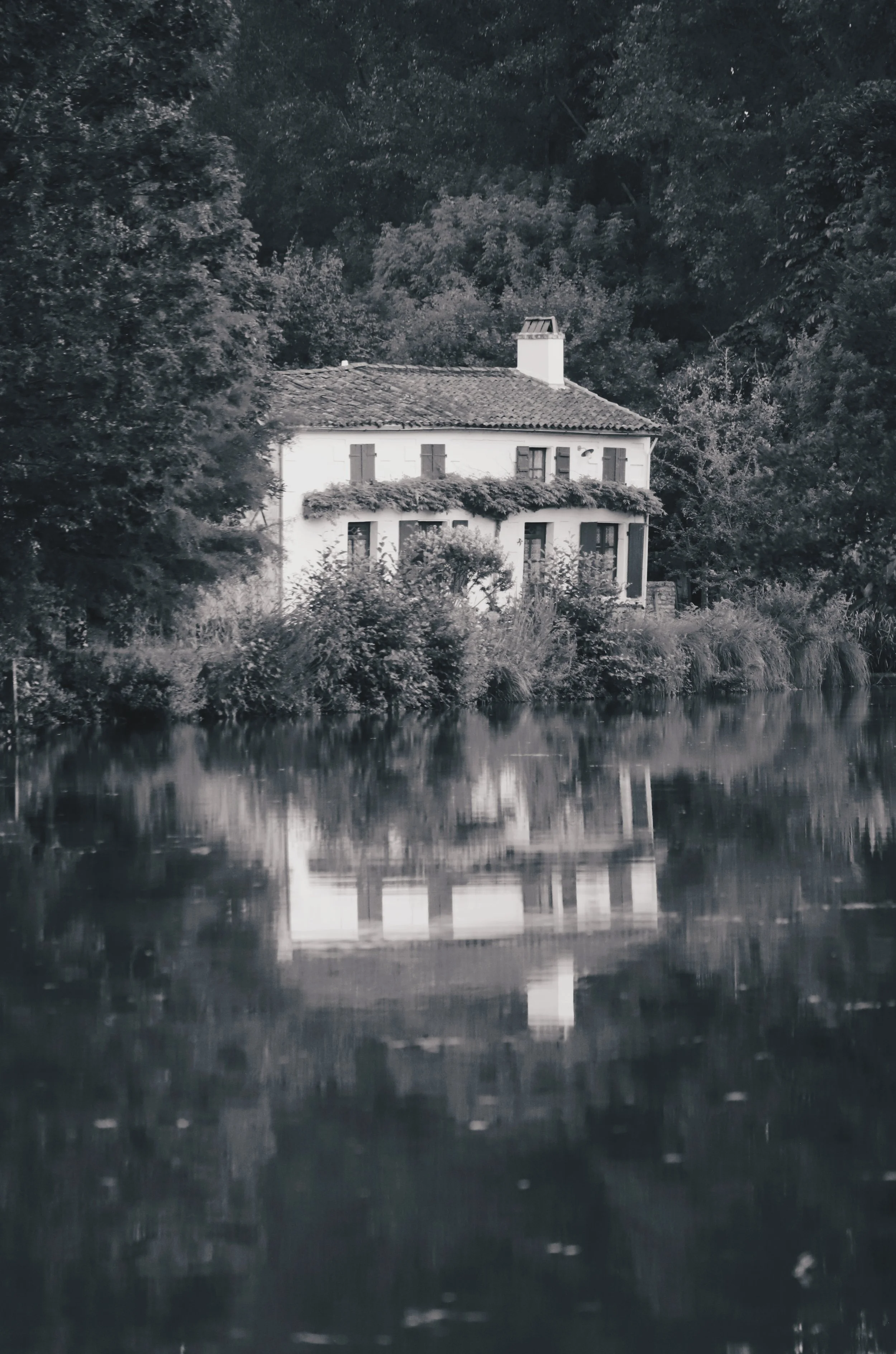 Une maison blanche à deux étages entourée d'arbres, située au bord d'une rivière, avec réflexion dans l'eau.