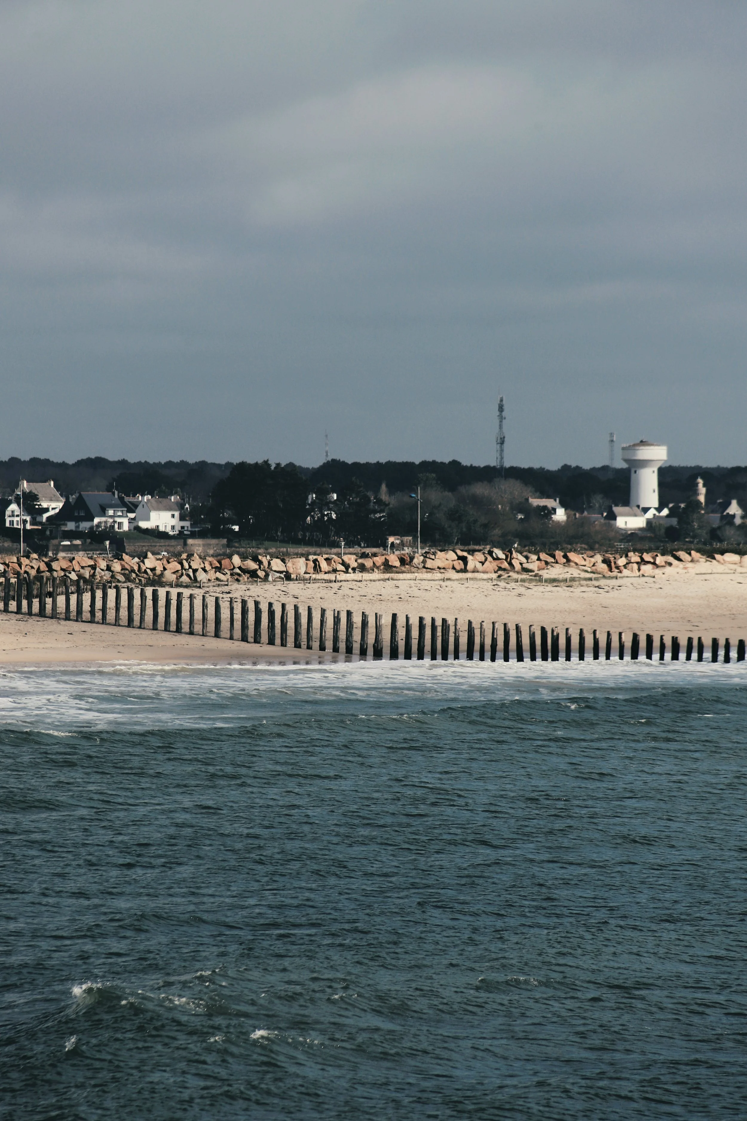 Une plage avec une digue en bois et une ville en arrière-plan, sous un ciel nuageux.