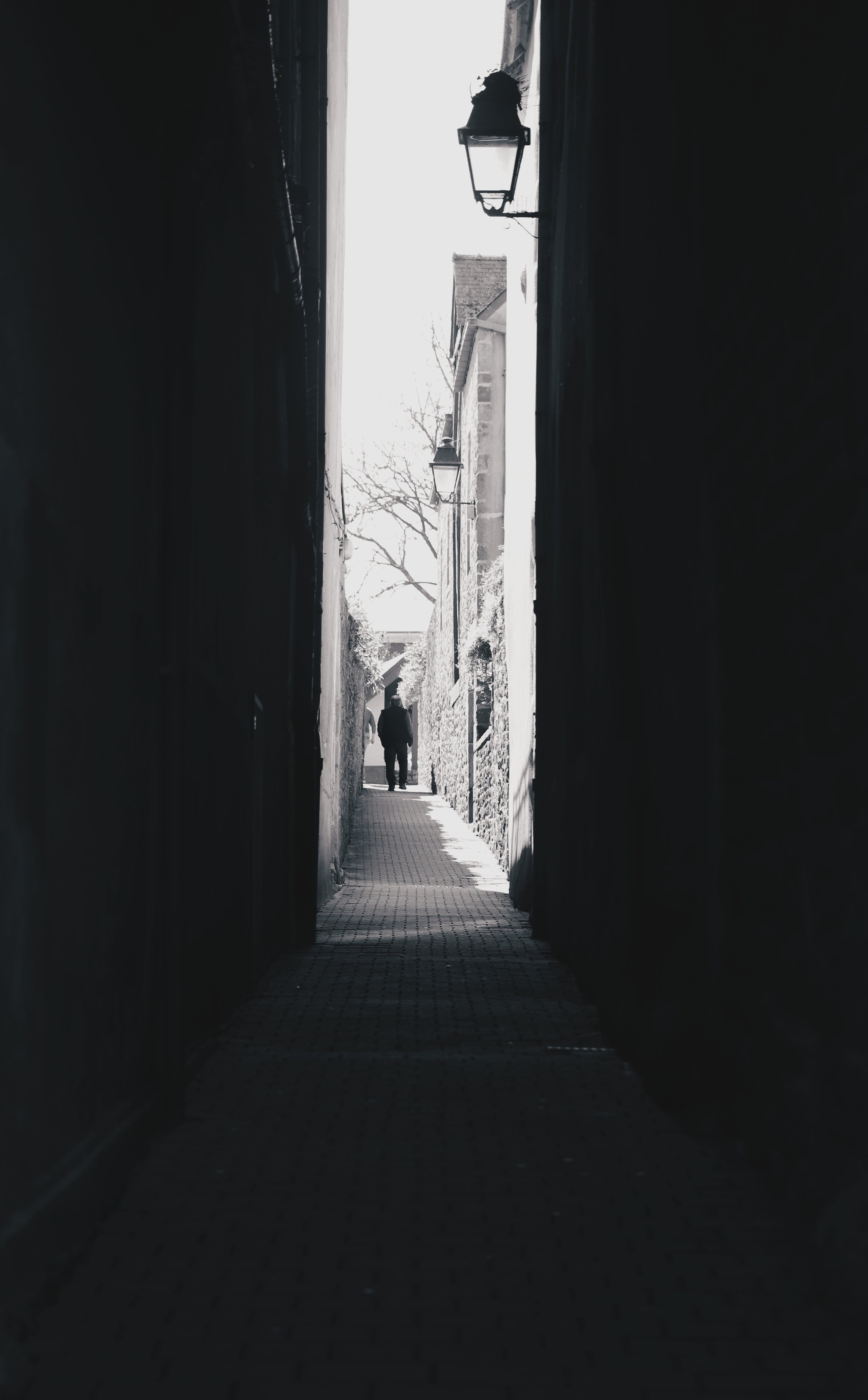 Vue d'une allée étroite entre des bâtiments, avec lampadaires accrochés aux murs, et deux personnes marchant au loin, en plein jour. L'image est en noir et blanc.
