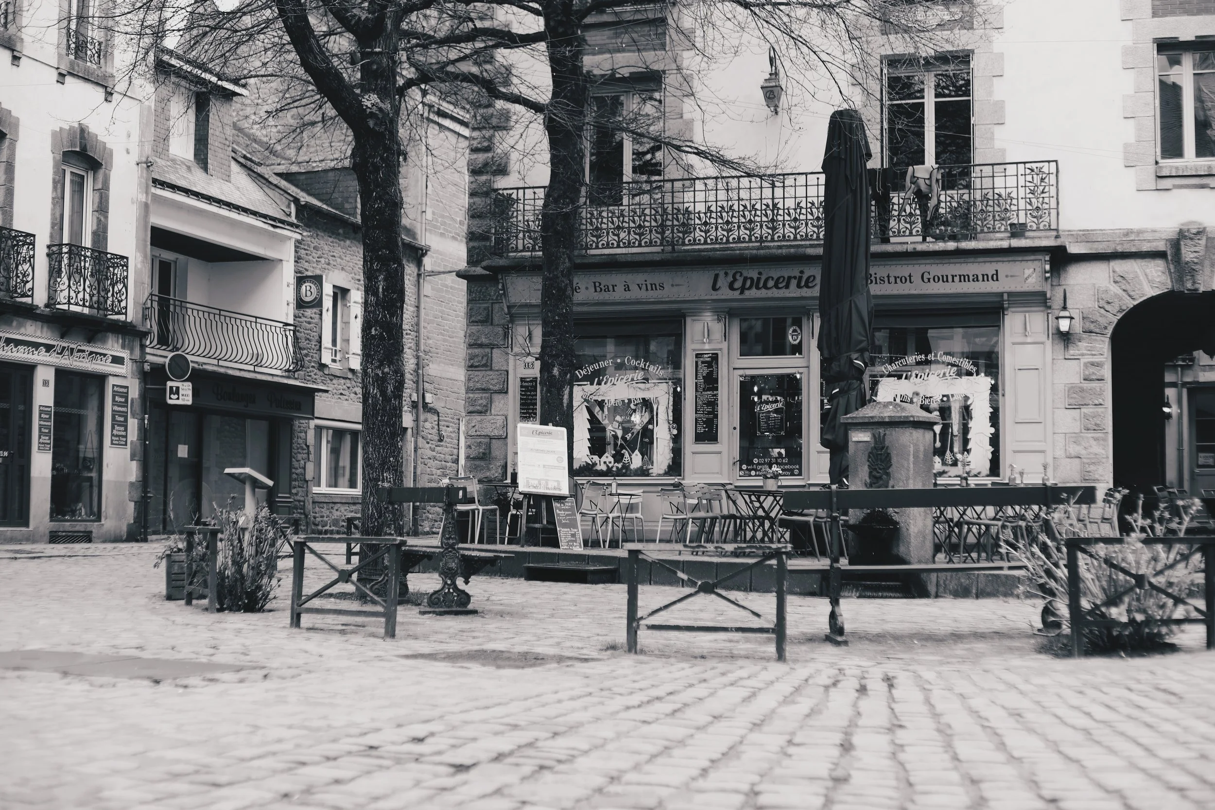 Une façade de bistrot avec des chaises et tables à l'extérieur, des arbres et un lampadaire dans une rue pavée, en noir et blanc.