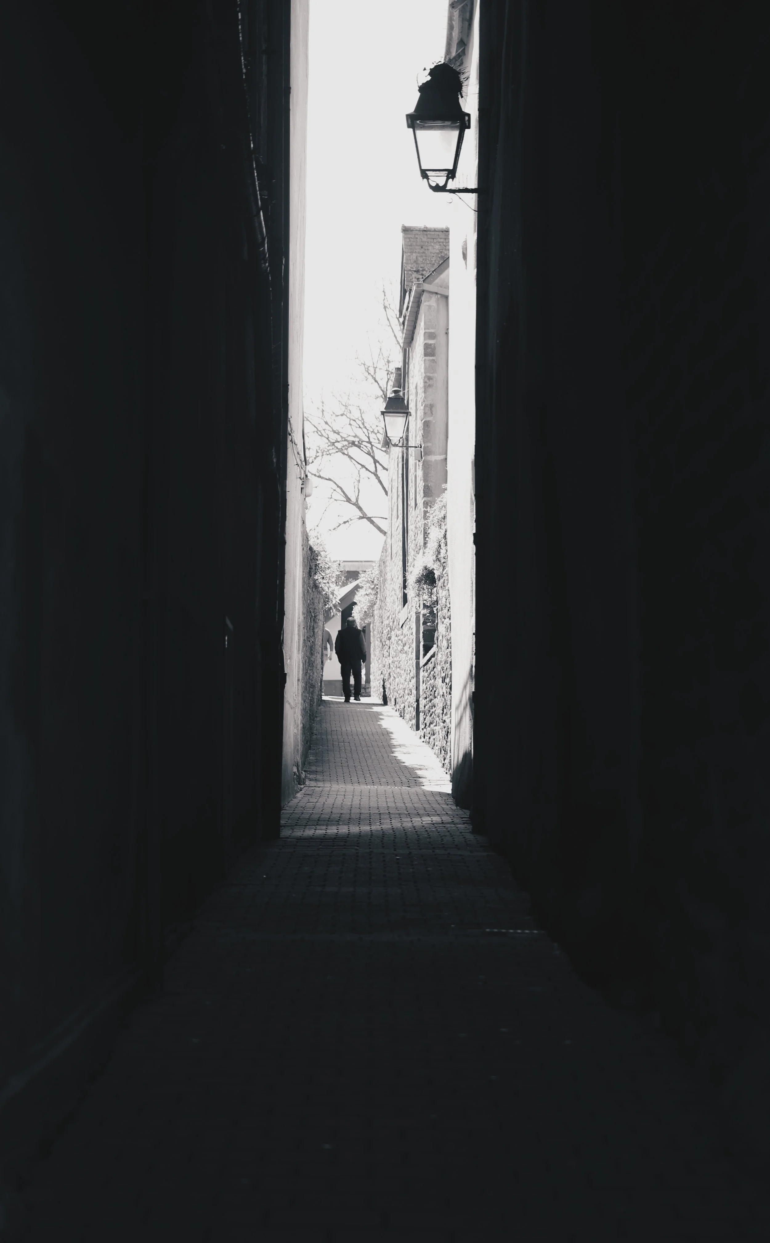 Vue d'une ruelle étroite avec des murs sombres, des lampadaires suspendus, et un homme marchant au loin sous un ciel clair.