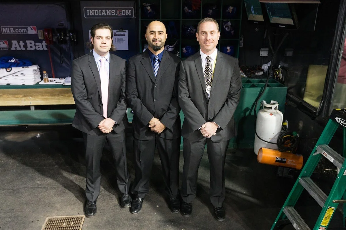 Three men in suits standing side by side indoors, with sports-themed signage and equipment visible in the background.
