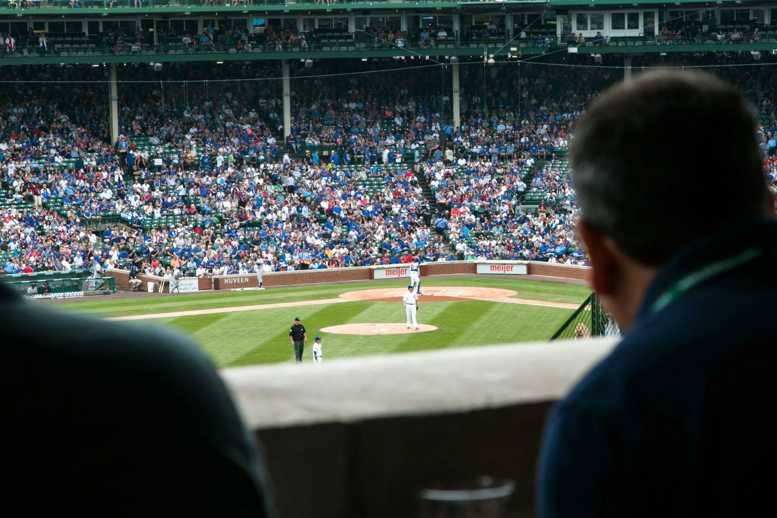 View of a baseball stadium from behind a person watching the game through a gap in the seats, with fans seated in the stands and players on the field.