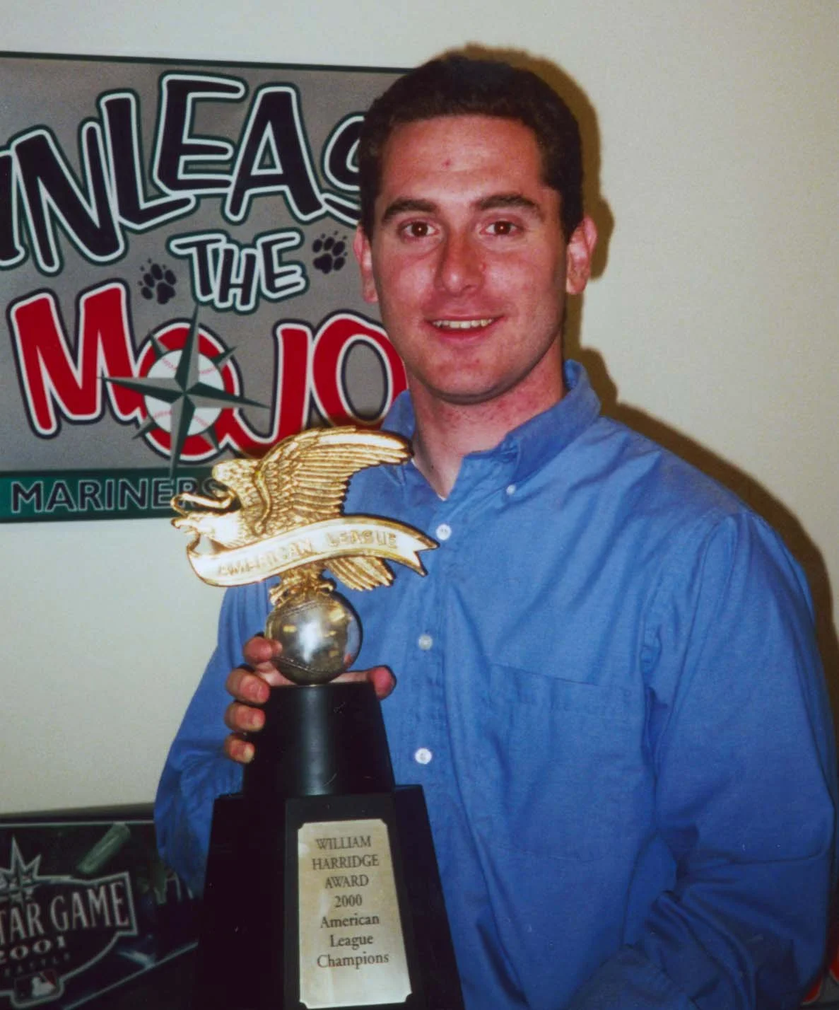 A young man in a blue shirt holding a trophy with a golden eagle on top. In the background, there is a sign that reads 'Rolling the Mo-Jo' and 'Mariners'.