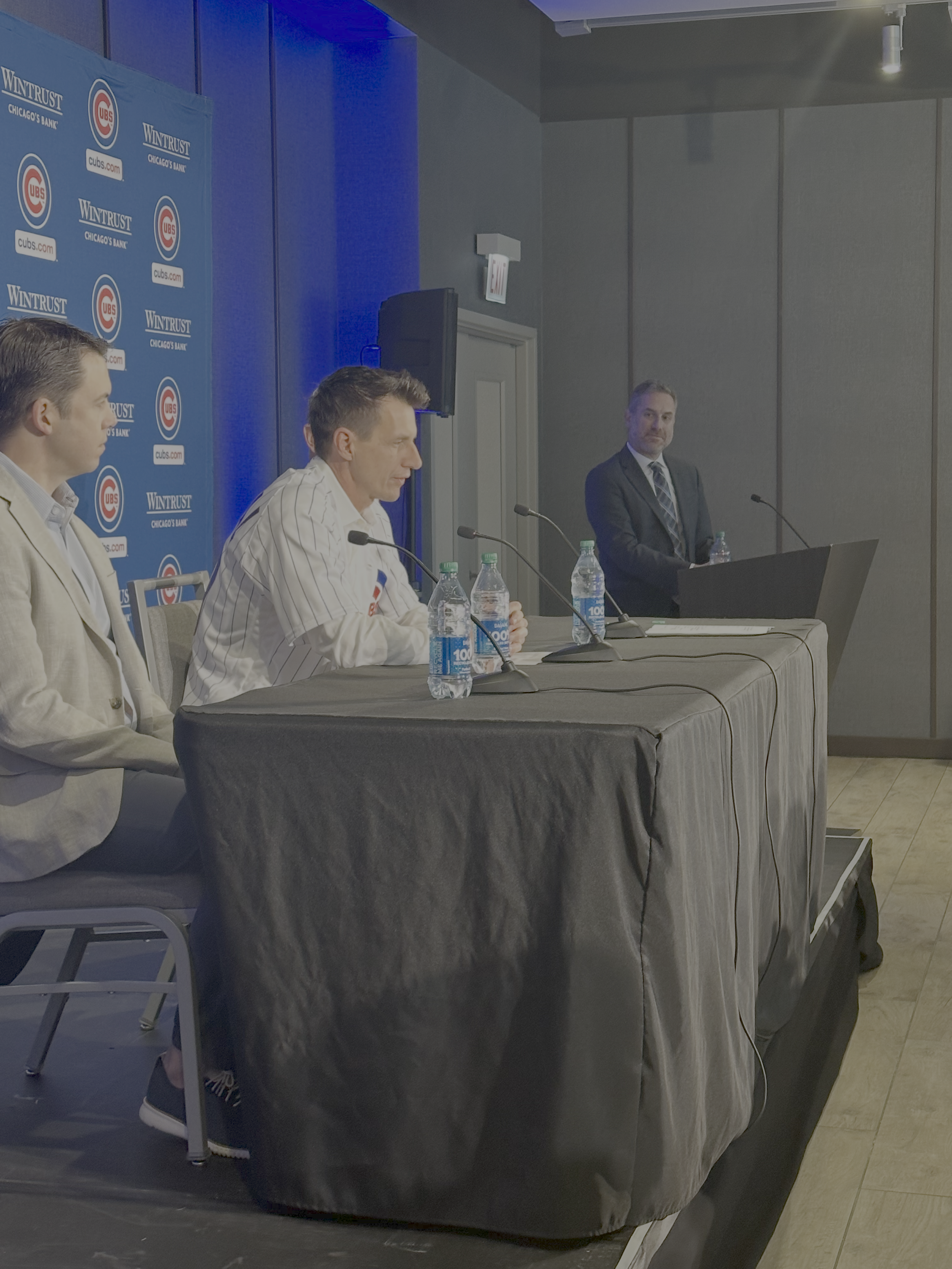 Press conference with four men sitting at a table with microphones and water bottles, a backdrop with Texas Rangers logos, and a man standing at a podium.