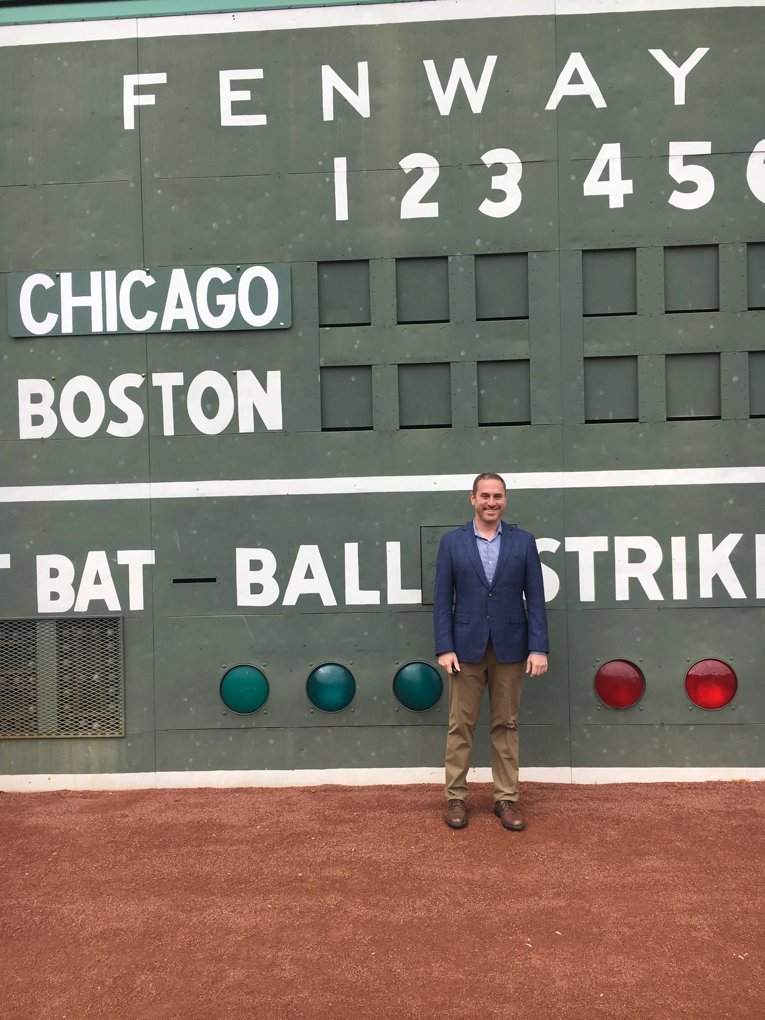 Man in blue blazer and tan pants standing on baseball field in front of green scoreboard with 'FENWAY 1 2 3 4 5 6' and 'CHICAGO BOSTON' names, baseball dugout and stadium lights.
