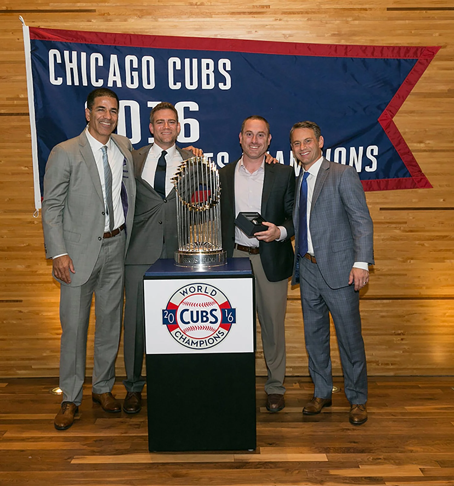 Four men in suits standing behind a podium with a Chicago Cubs championship banner and trophy, celebrating the Cubs' 2016 World Series win.