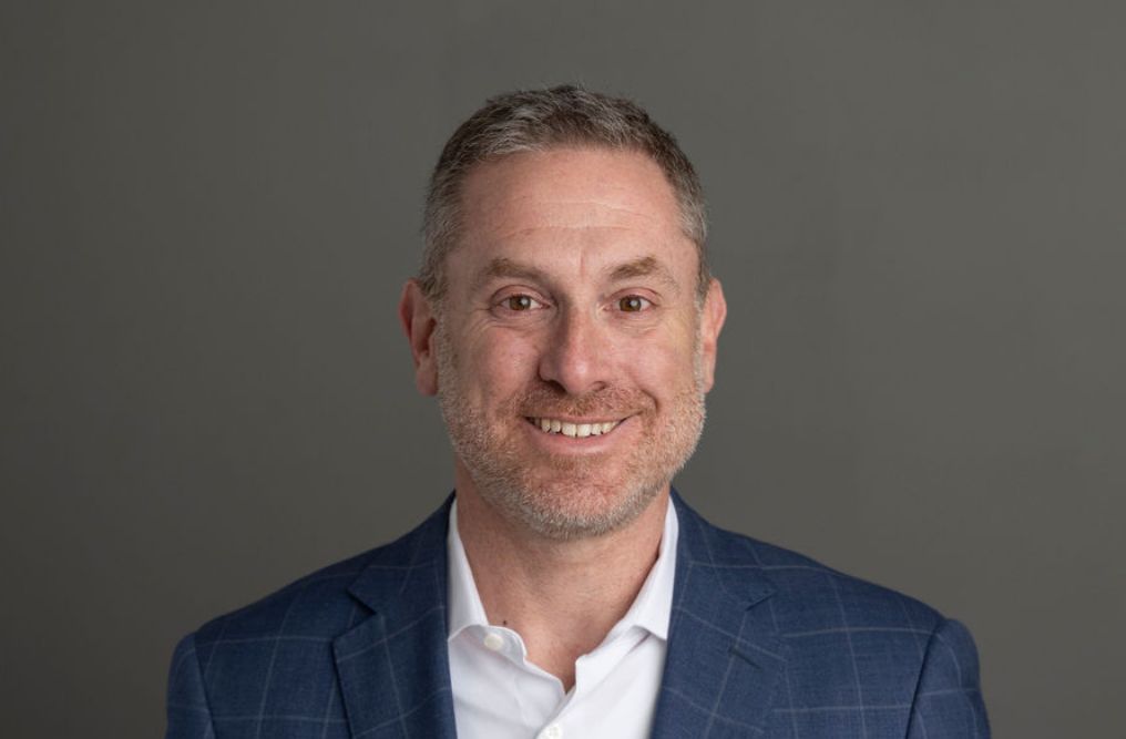 Headshot of a smiling man with short gray hair and beard, wearing a dark blue checkered blazer and white shirt, against a plain gray background.