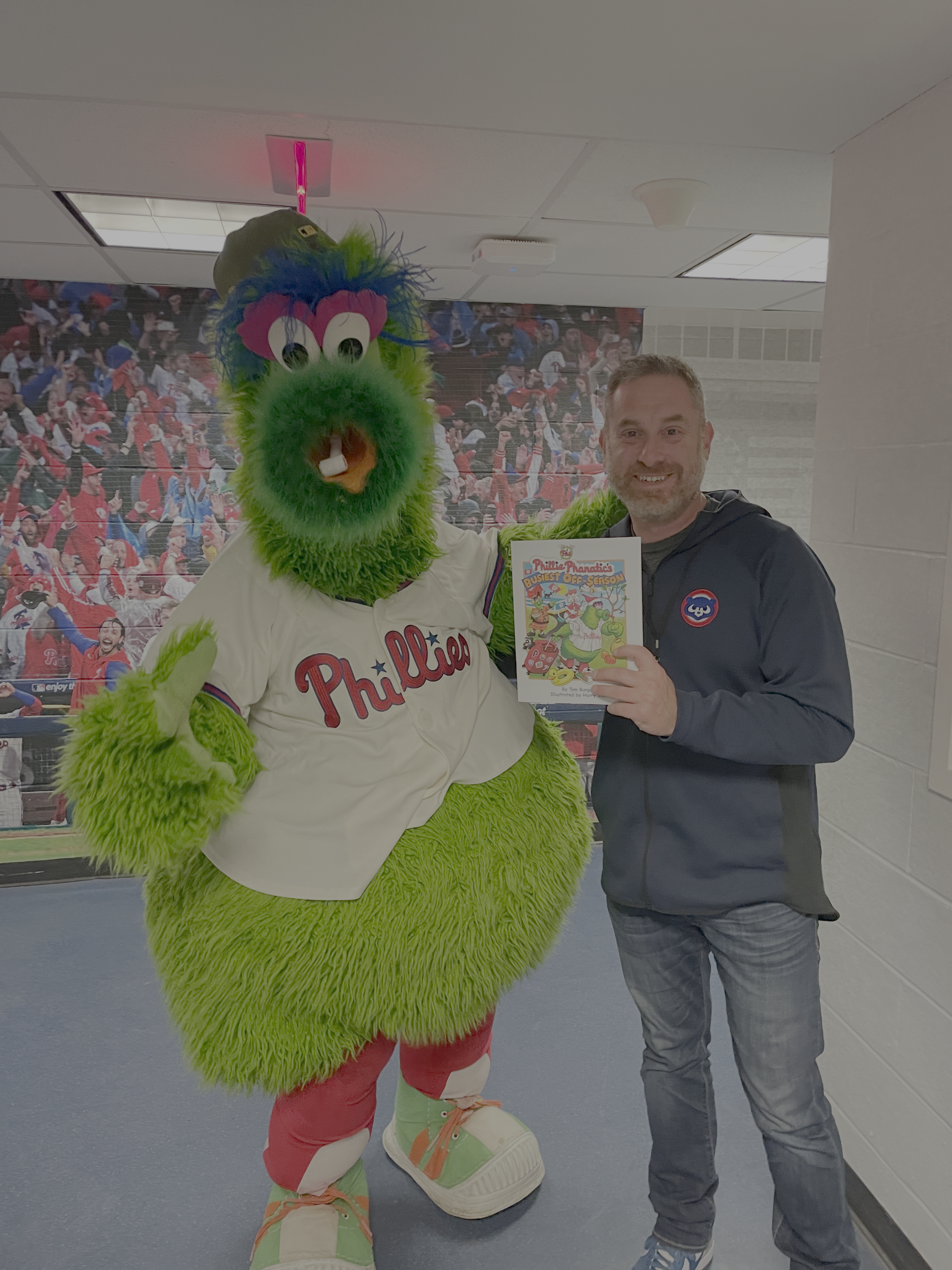 A man standing next to a person in a Dr. Seuss-themed costume, holding a book at an indoor event with a crowd in the background.