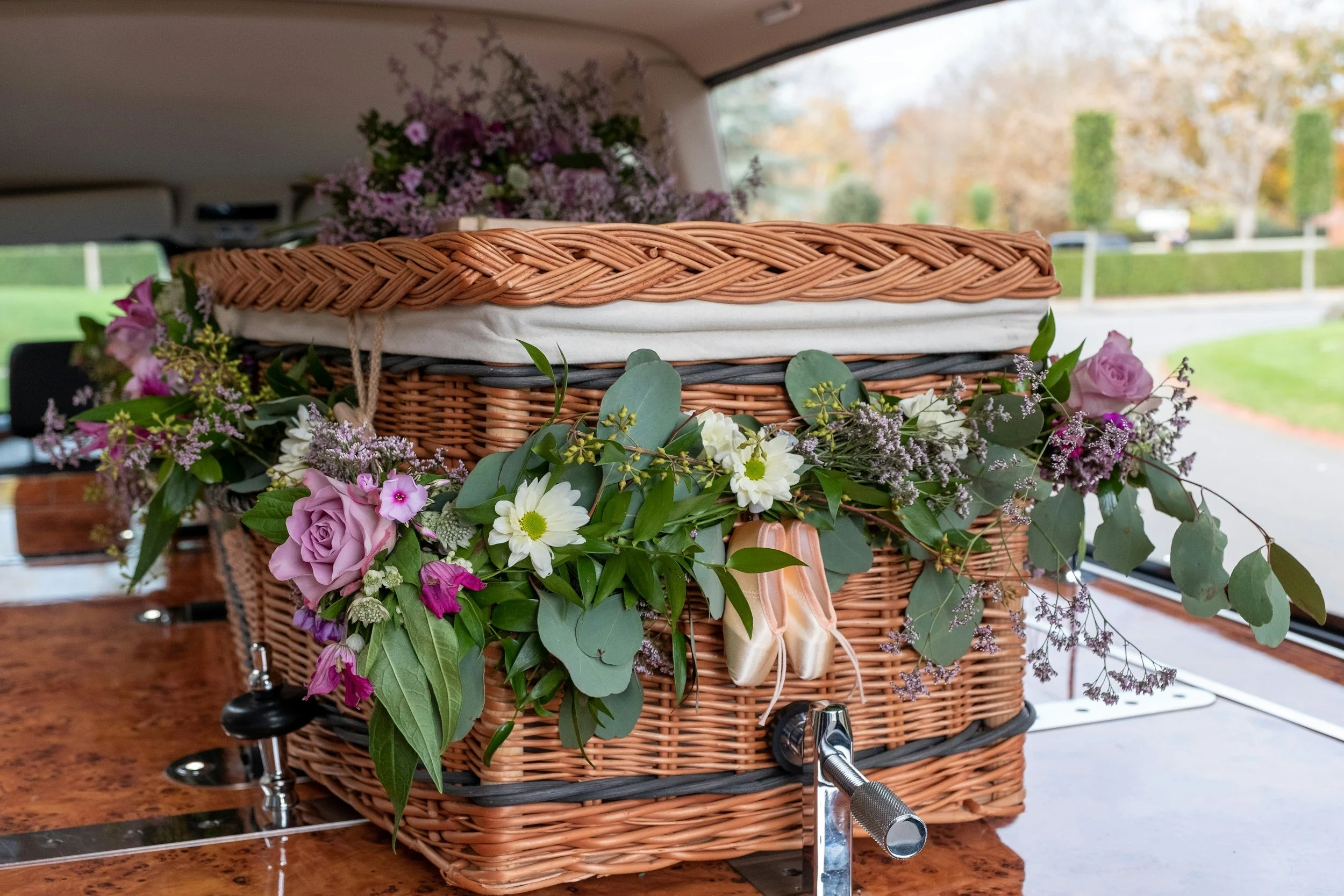 A wicker casket decorated with purple and white flowers inside a vehicle, with trees and a road visible outside the window.