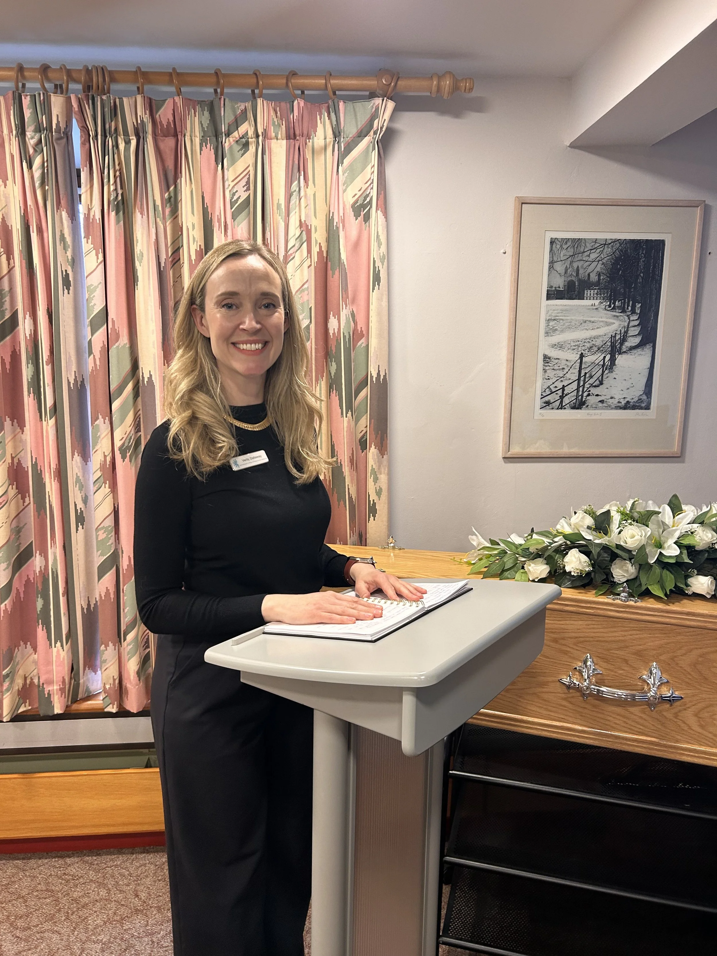 A woman standing at a podium, smiling, with a book open in front of her. She has long blonde hair and is wearing a black outfit. There are pink and green curtains in the background, with a framed black-and-white photograph of a snowy landscape on the wall behind her. A bouquet of white flowers is placed on a wooden surface beside her.