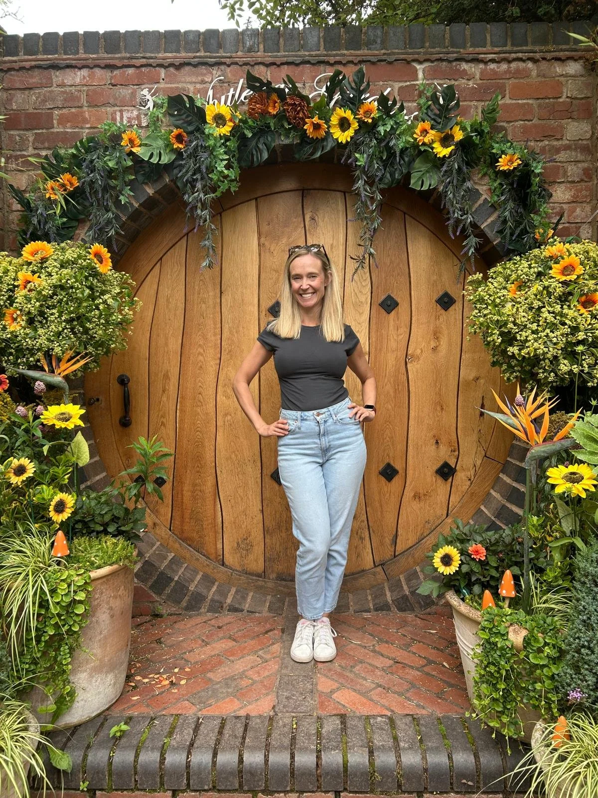 A woman with blonde hair wearing a black t-shirt, light blue jeans, and white shoes, standing in front of a large round wooden door decorated with flowers and greenery, outdoors with brick paving.
