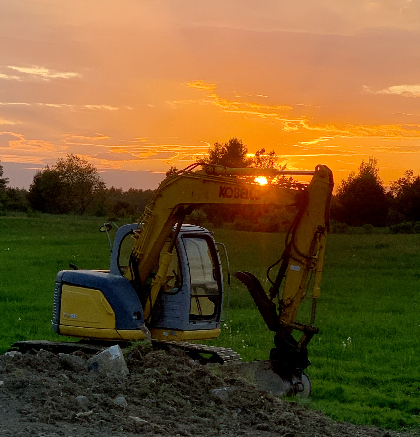 A yellow and blue excavator on a grassy field during sunset with an orange sky and scattered clouds, surrounded by trees.