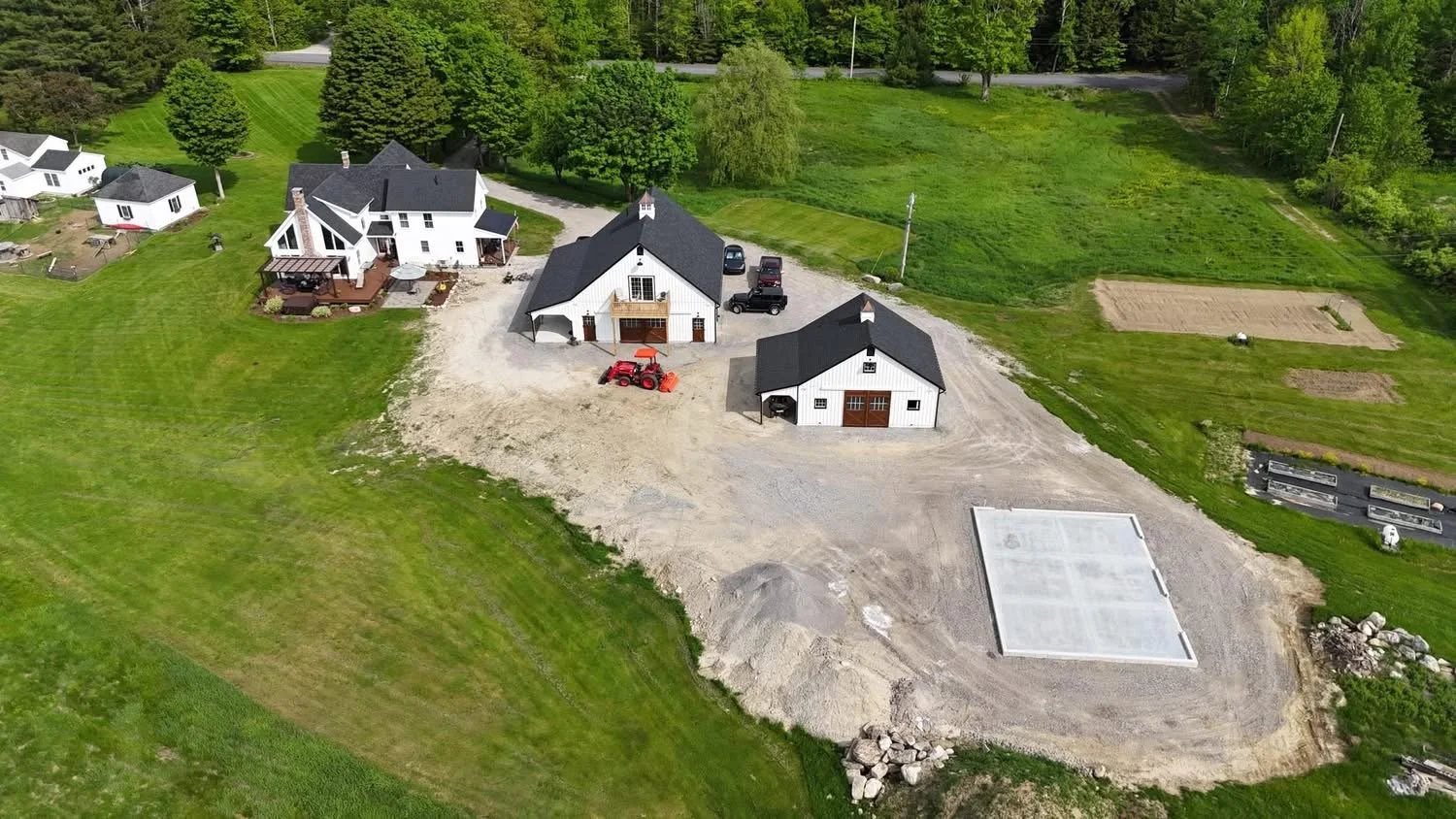 Aerial view of a rural property with a house, a barn, a garage under construction, parked vehicles, and surrounding greenery.