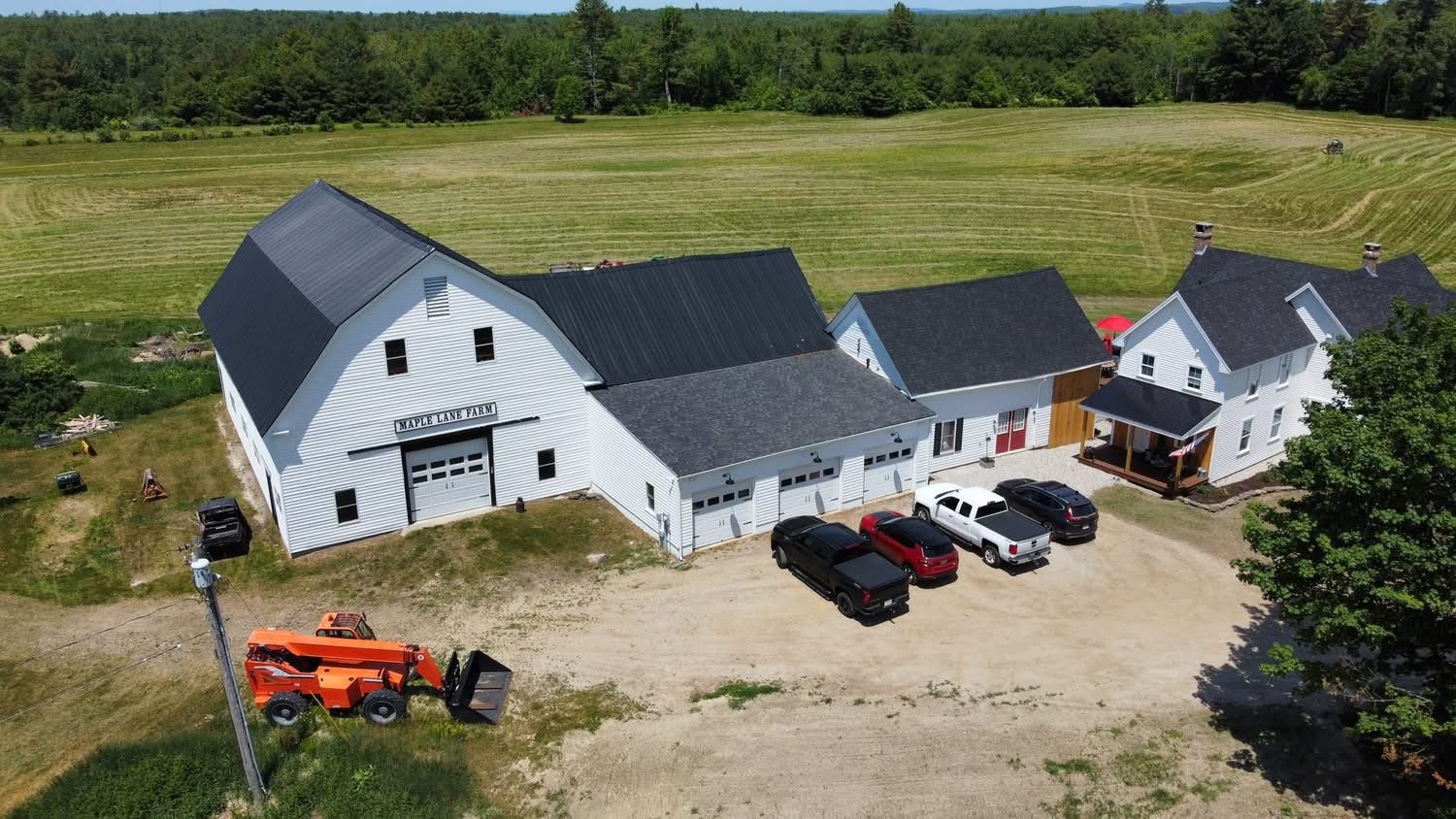 Aerial view of a white farm building labeled 'Maple Lane Farm' with a black metal roof, surrounded by a dirt parking area with several parked cars, a tractor, and a large open field with trees in the background.