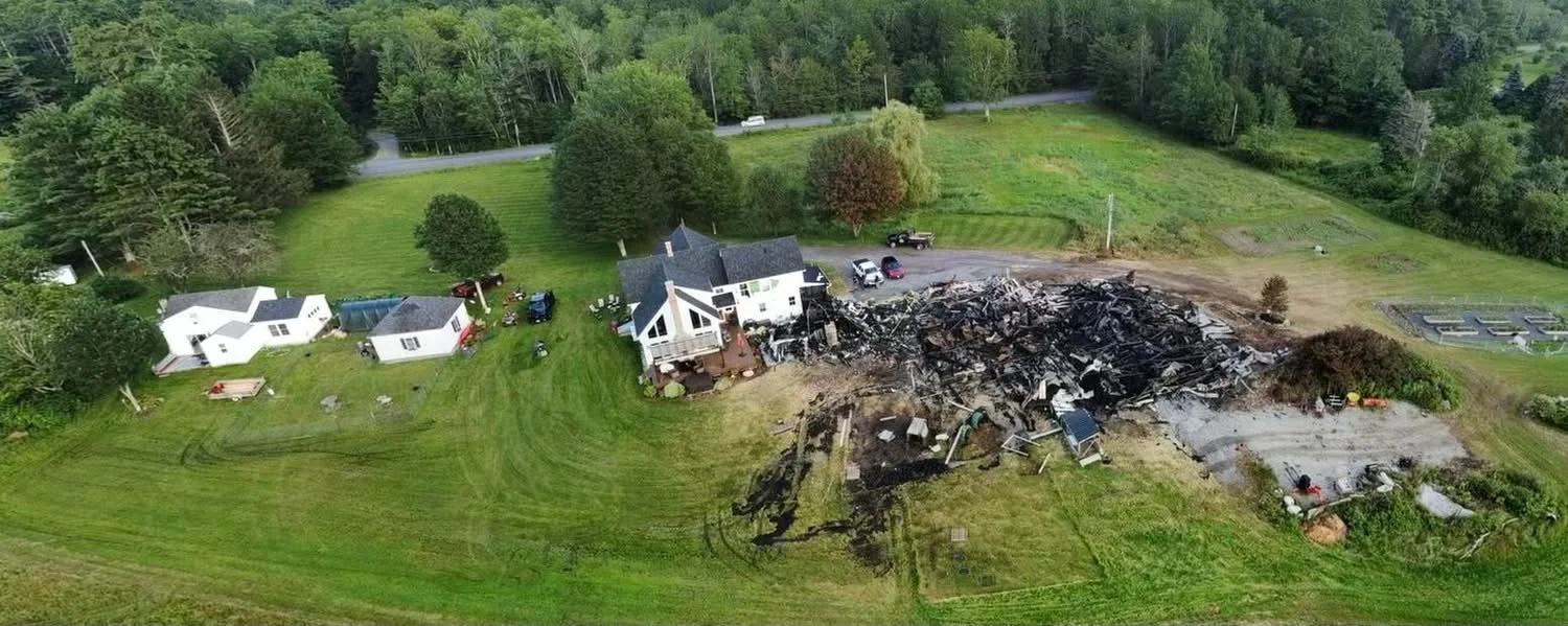 Aerial view of a damaged house and burned debris in a yard with green grass and trees.