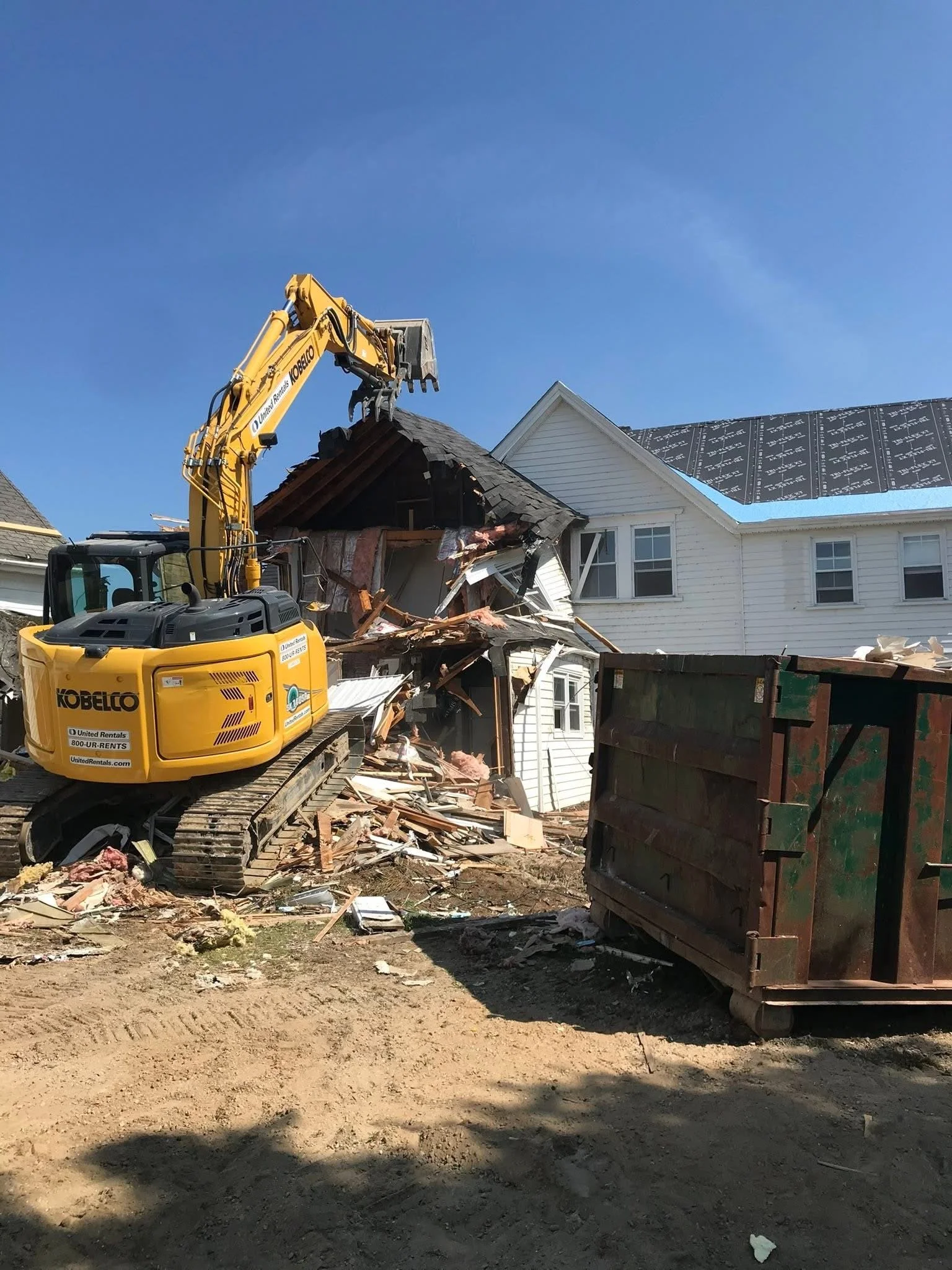 A construction scene showing a yellow excavator demolishing the side of a house, with debris scattered around and a large dumpster near the house.