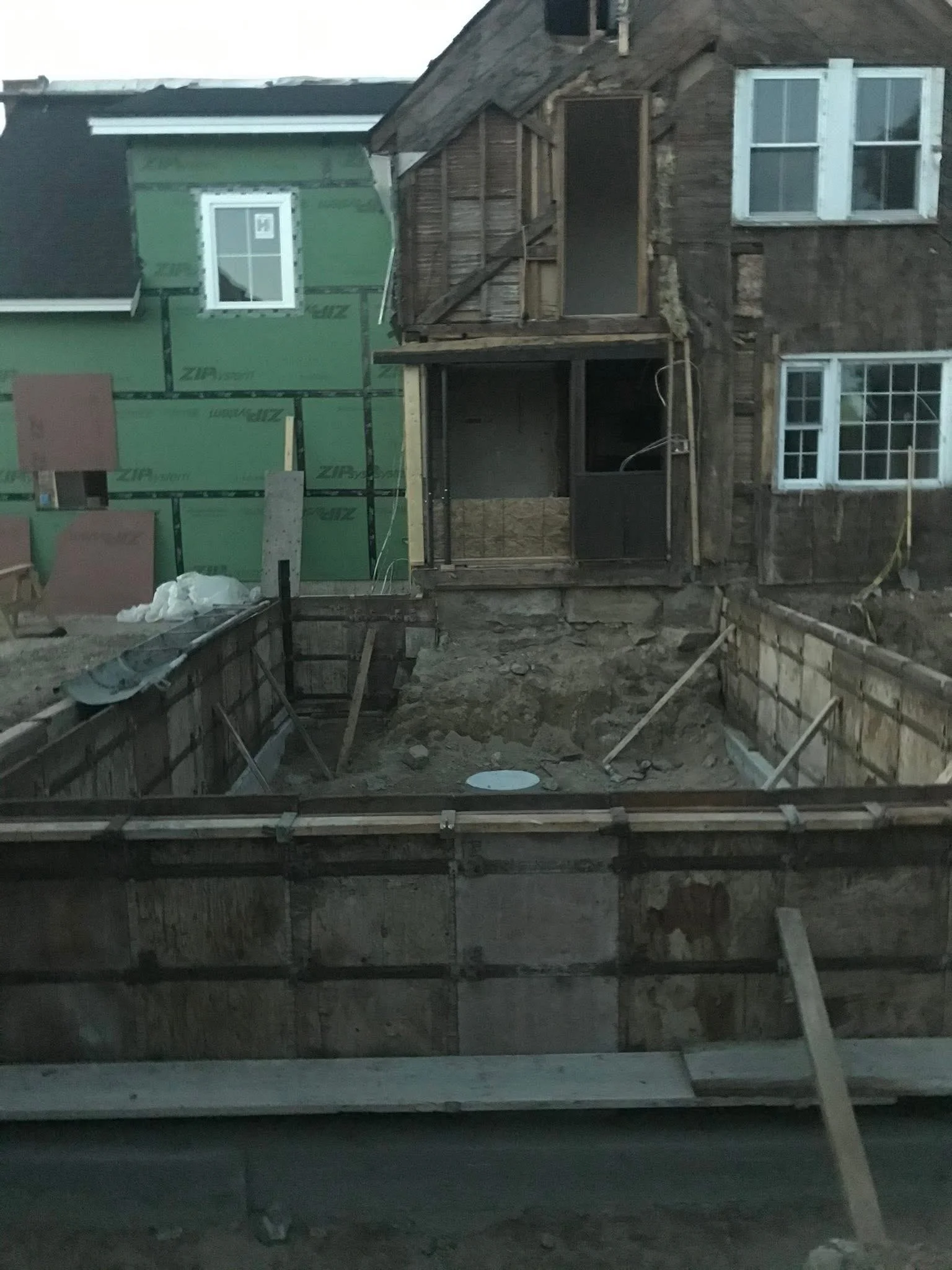 Construction site with a partially demolished house and an open foundation trench. The house has weathered wooden walls and several windows, some with white frames. On the left, there is green insulation or weather barrier on part of the building. Building materials and tools are visible around the site.